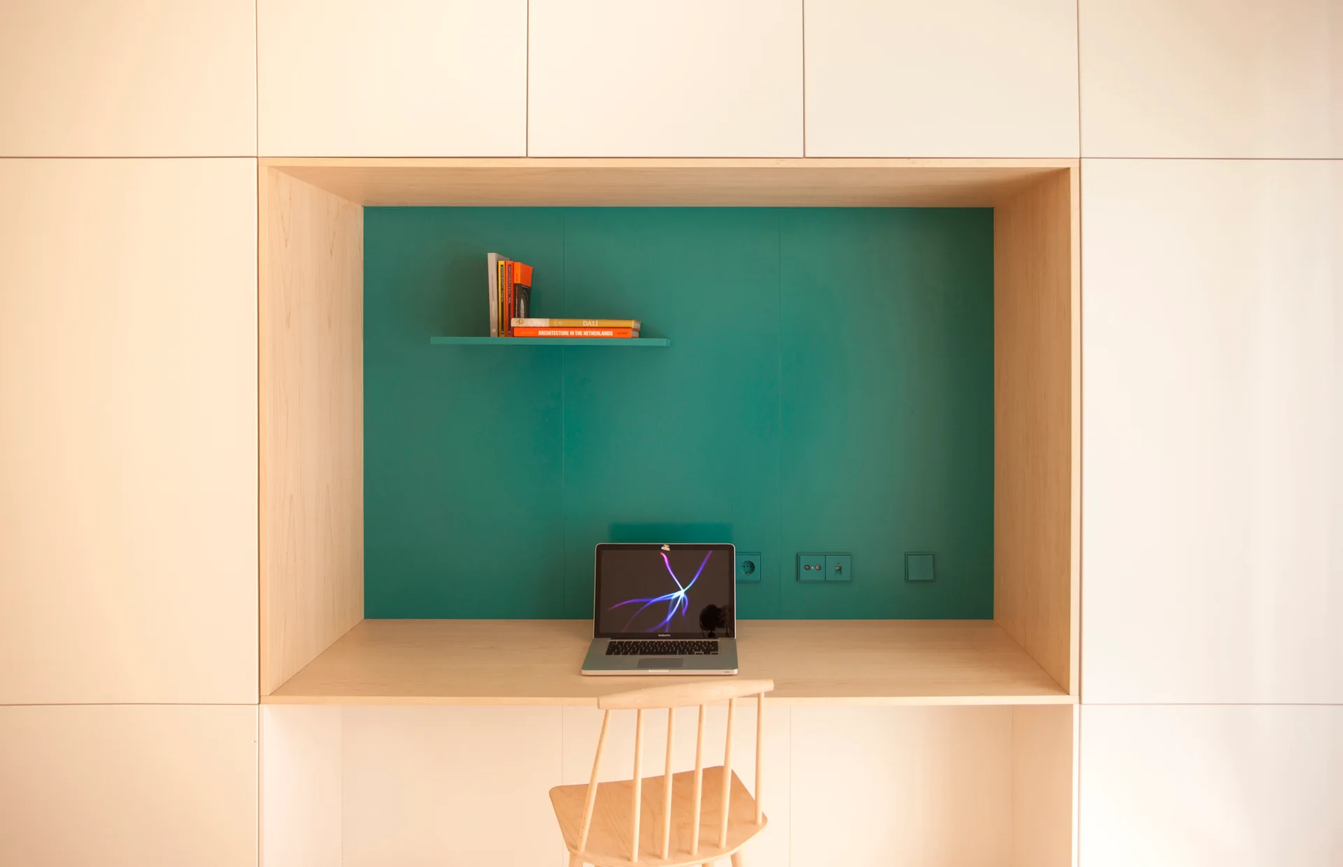 A light wood built-in desk recessed into matte white cabinets, featuring a bold teal accent wall, a floating shelf, and a wooden chair.