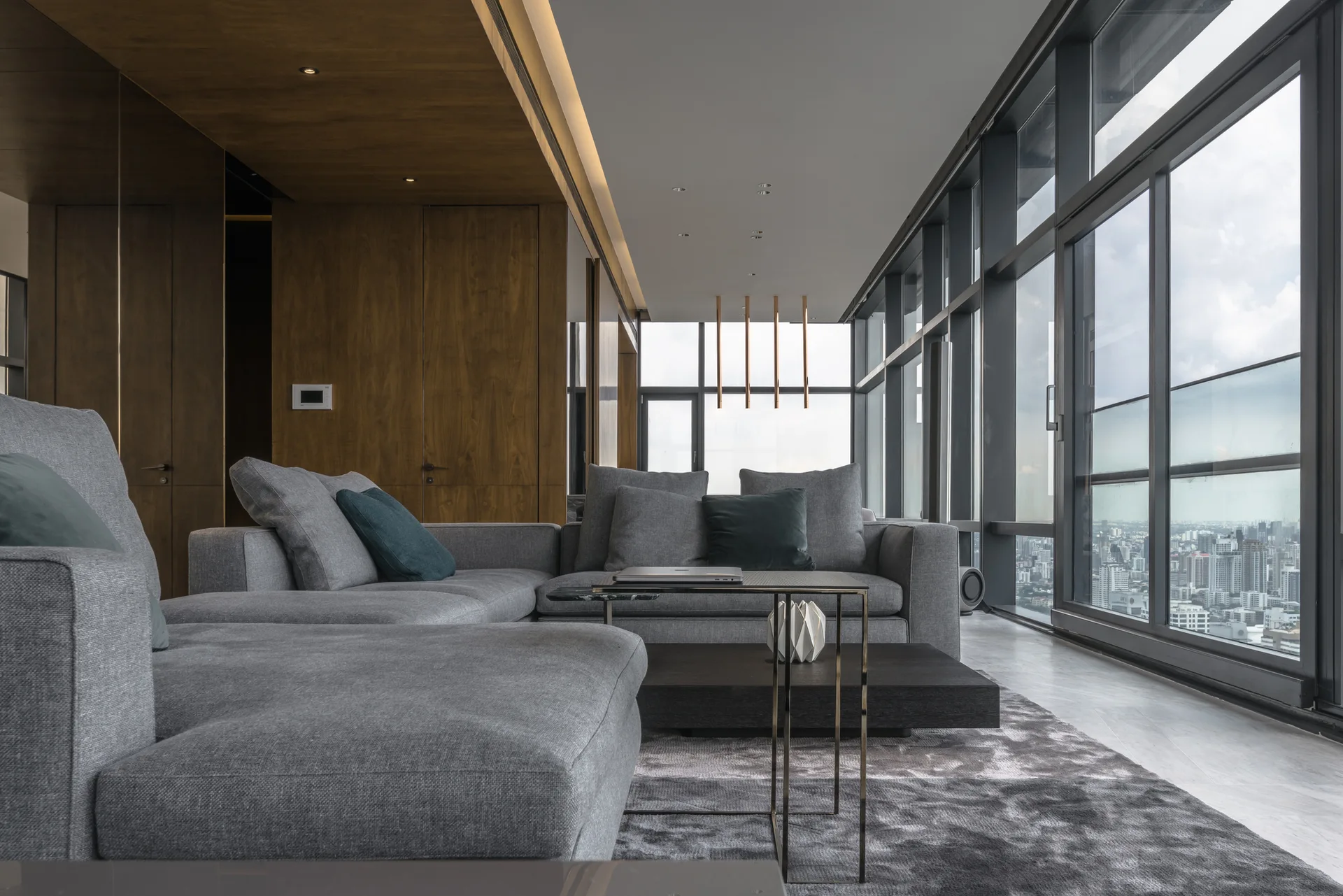 A grey sectional sofa sits on a textured rug in a living room featuring floor-to-ceiling windows and a warm wood-paneled accent wall.