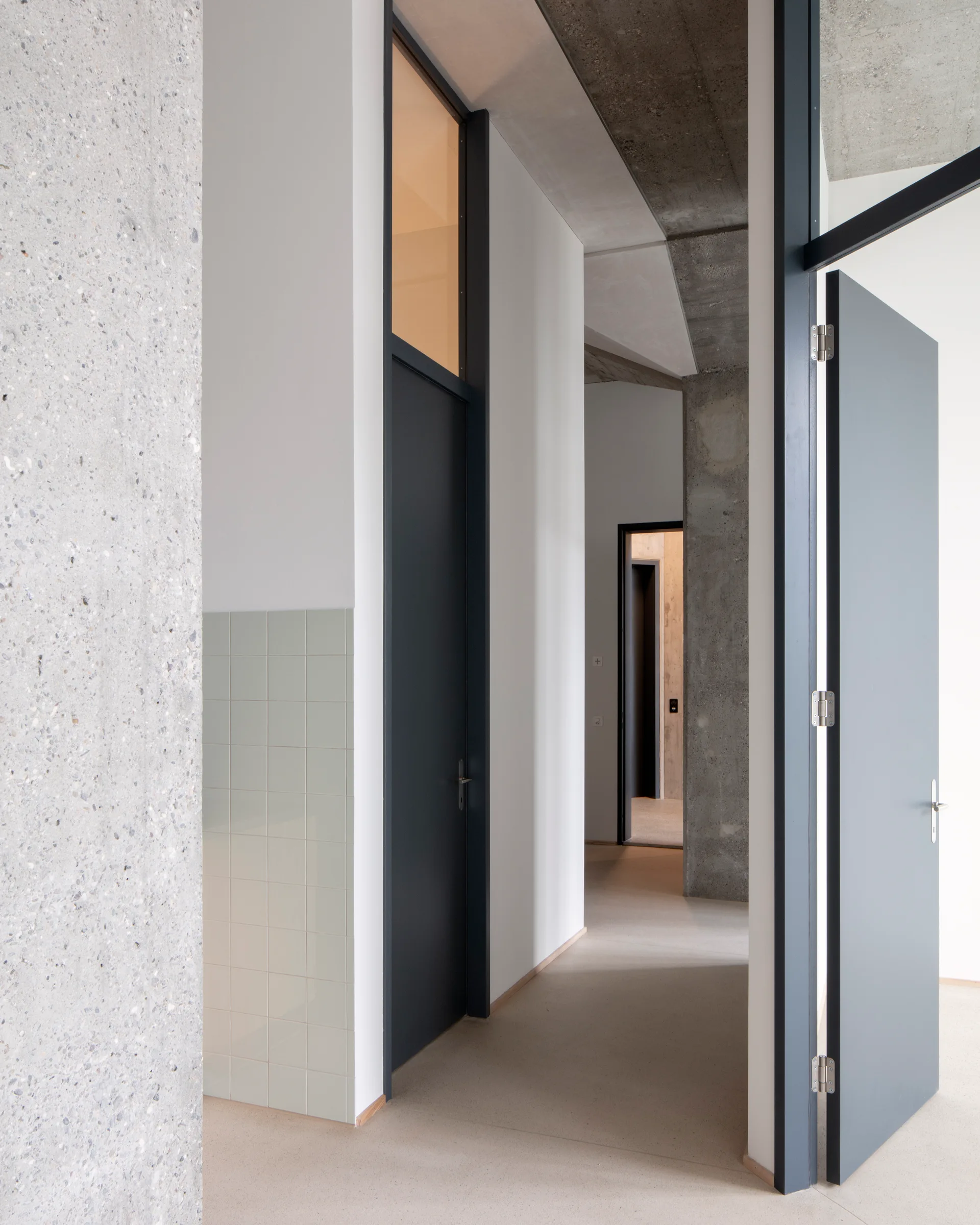 A minimalist hallway features dark grey doors, a raw exposed concrete column, and a small section of light green square wall tiles.