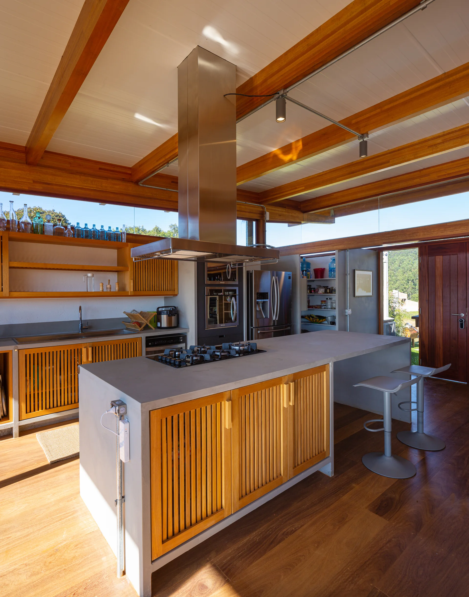 A modern kitchen features a concrete-topped island with slatted wooden cabinet doors, exposed ceiling beams, and a stainless steel range hood.