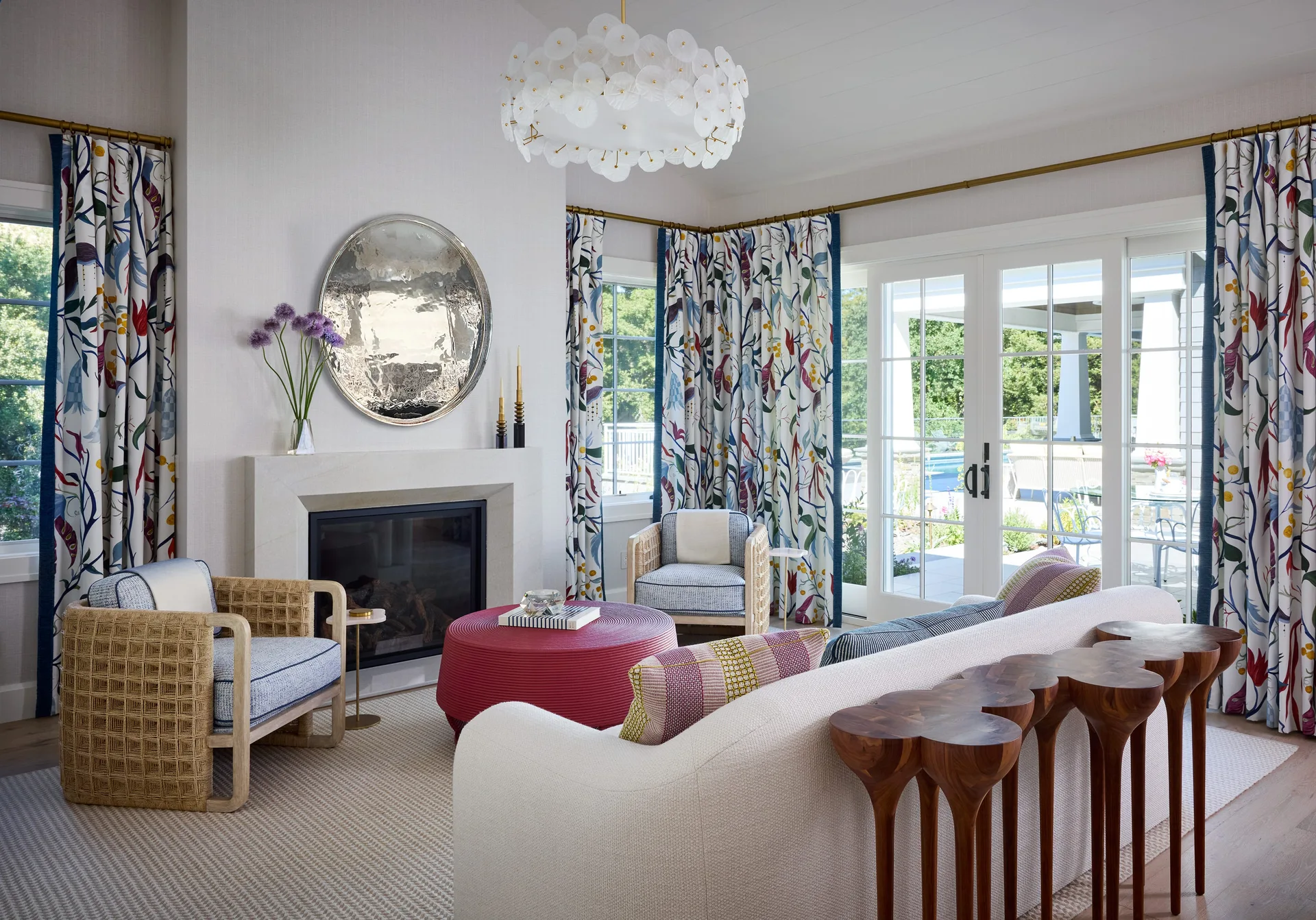 A bright living room featuring a stone fireplace, patterned floral curtains, a cream sofa, and a textured red ottoman.