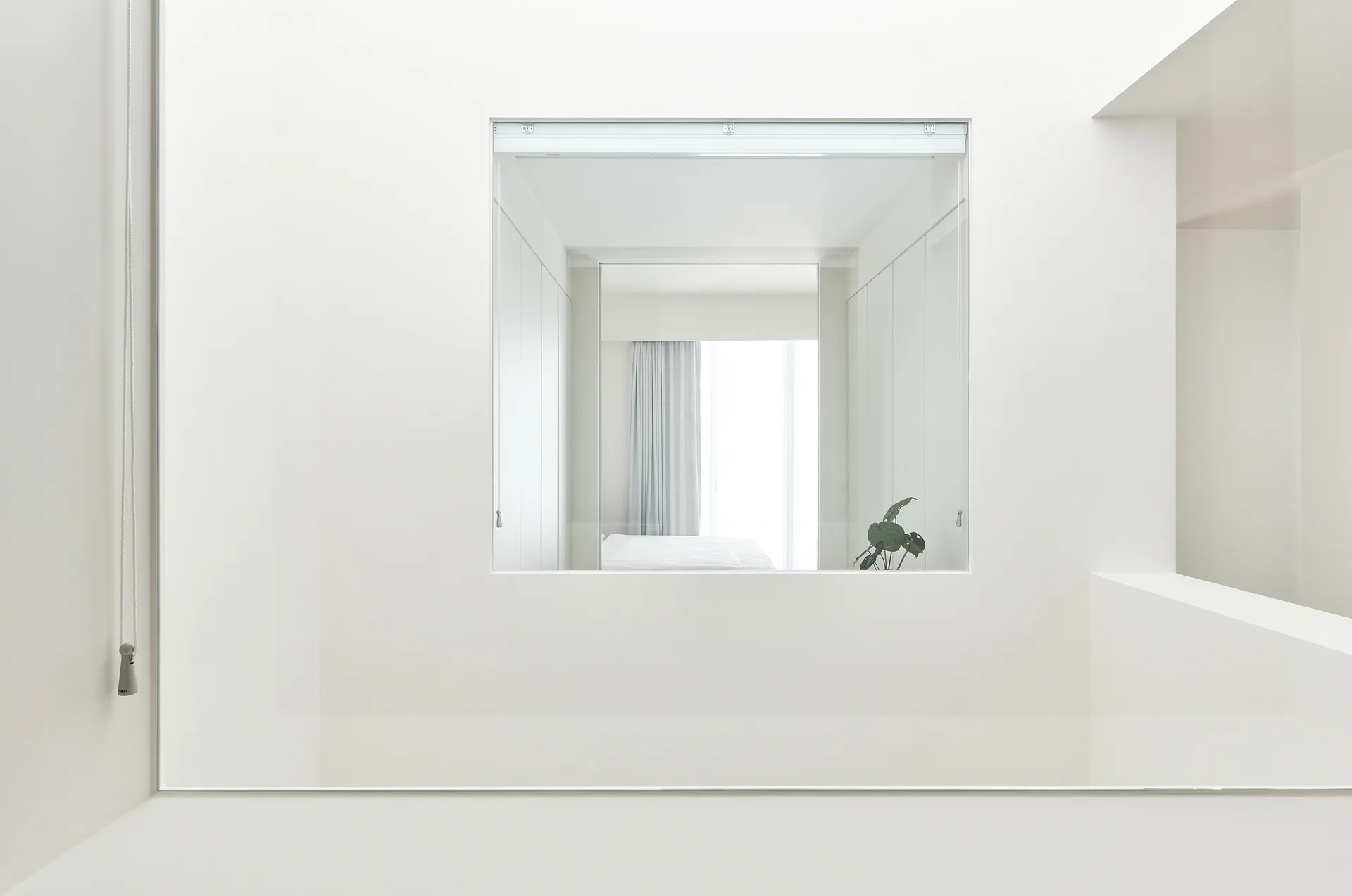 A minimalist white room with a square wall cutout framing a view of a clean bedroom with a bed and a green plant.