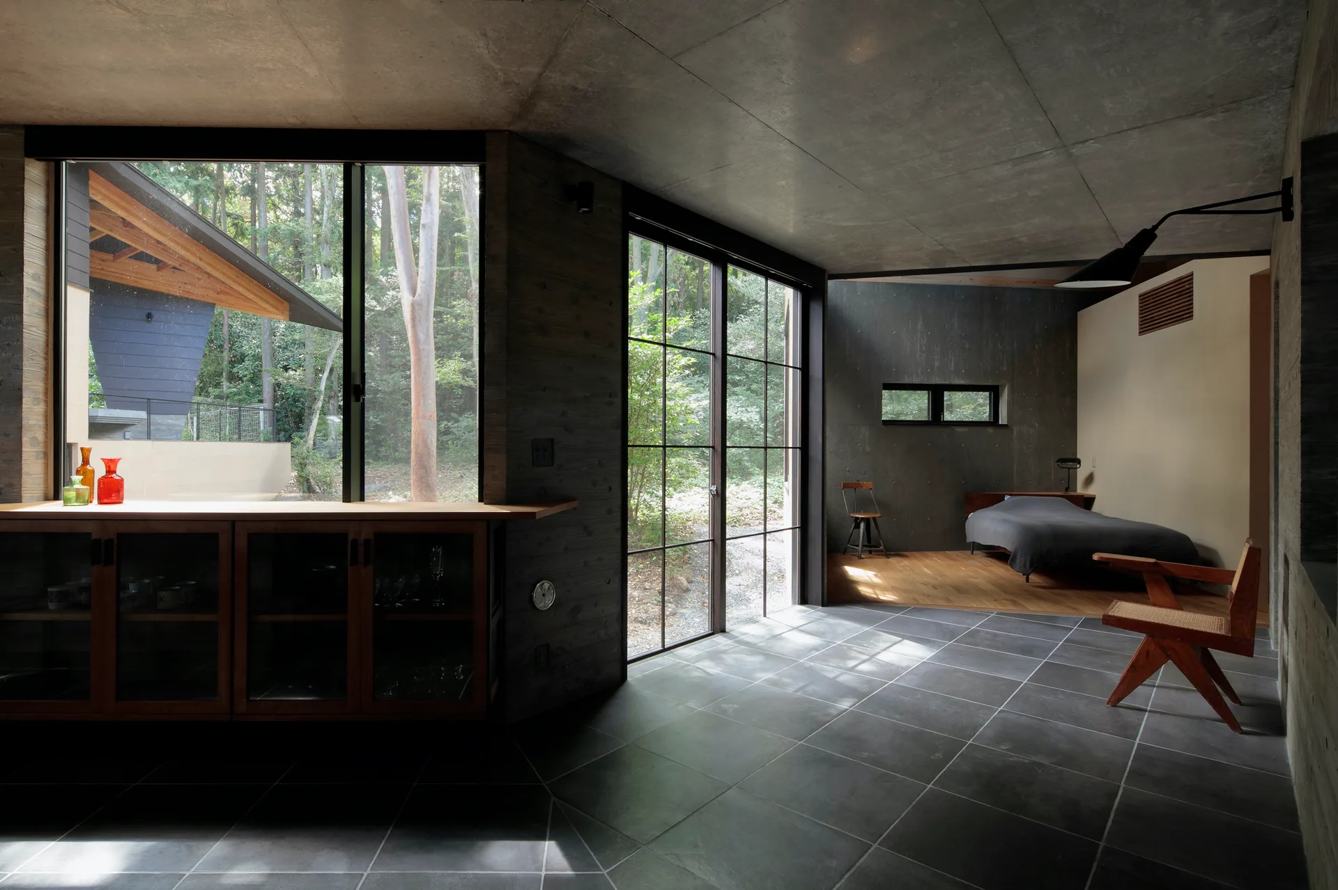 A modern bedroom with board-formed concrete walls, large glass windows facing a forest, and a low platform bed on a warm wood floor.