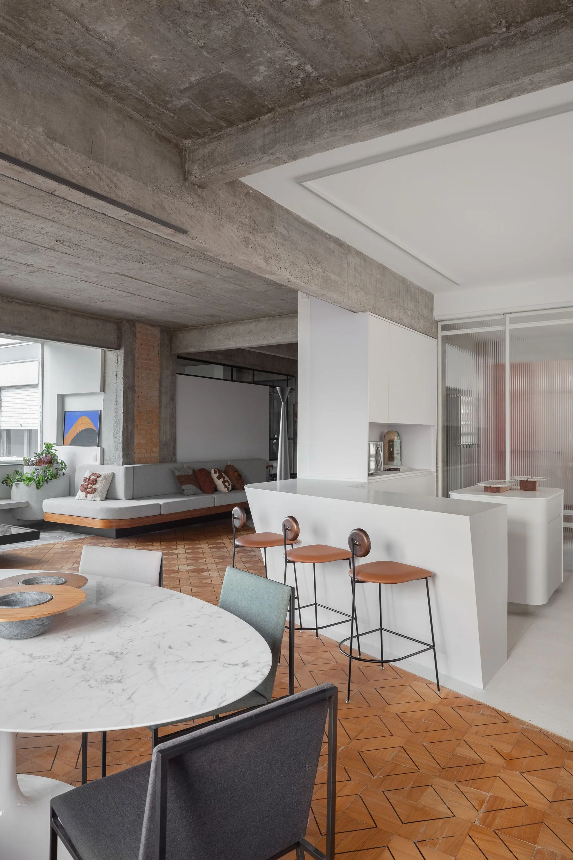 An open-concept living space features an exposed concrete ceiling, a white kitchen island with leather bar stools, and geometric parquet wood flooring.