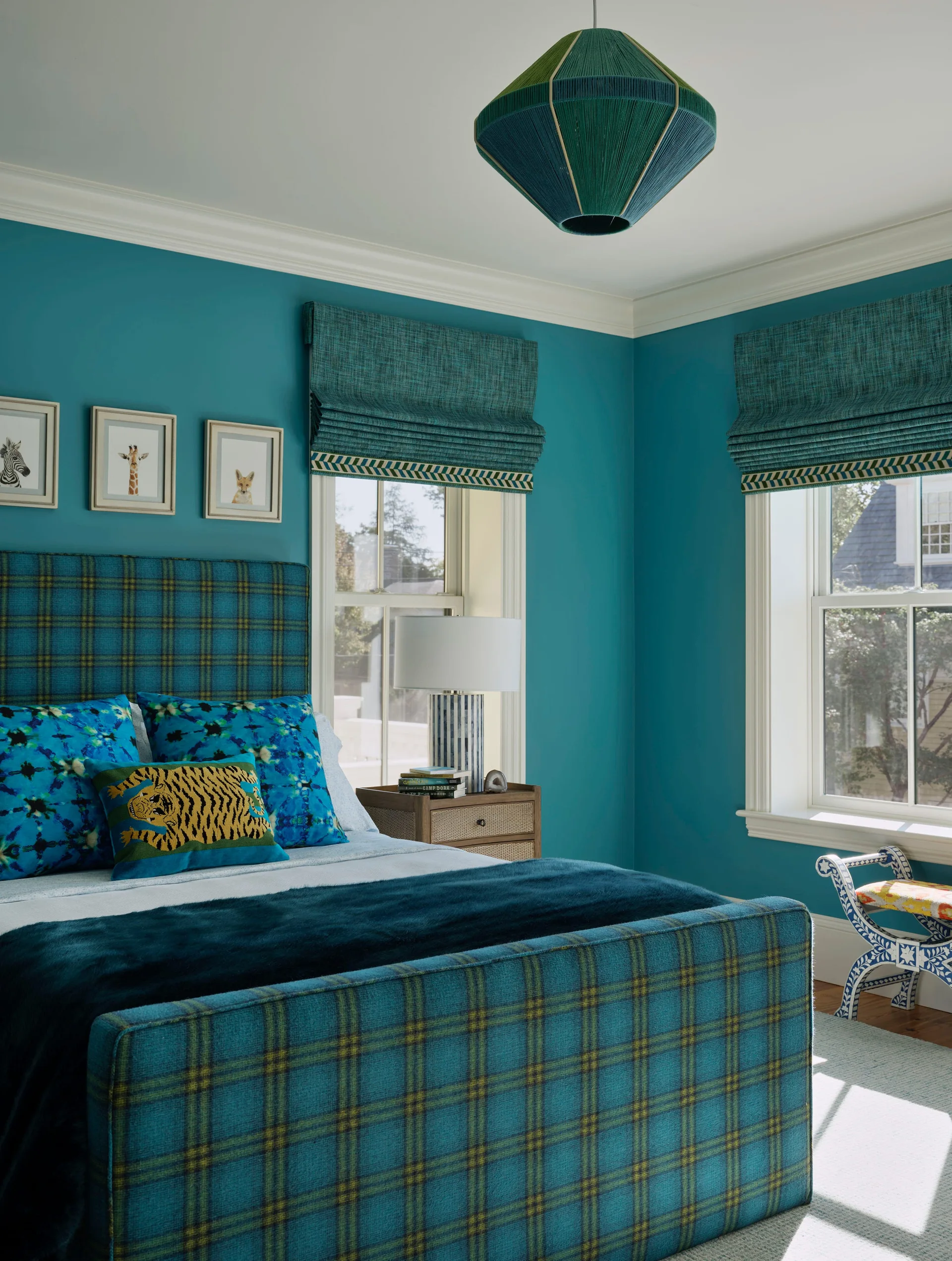 A teal bedroom features a plaid upholstered bed, matching teal roman window shades, three small framed animal prints, and a geometric pendant light.