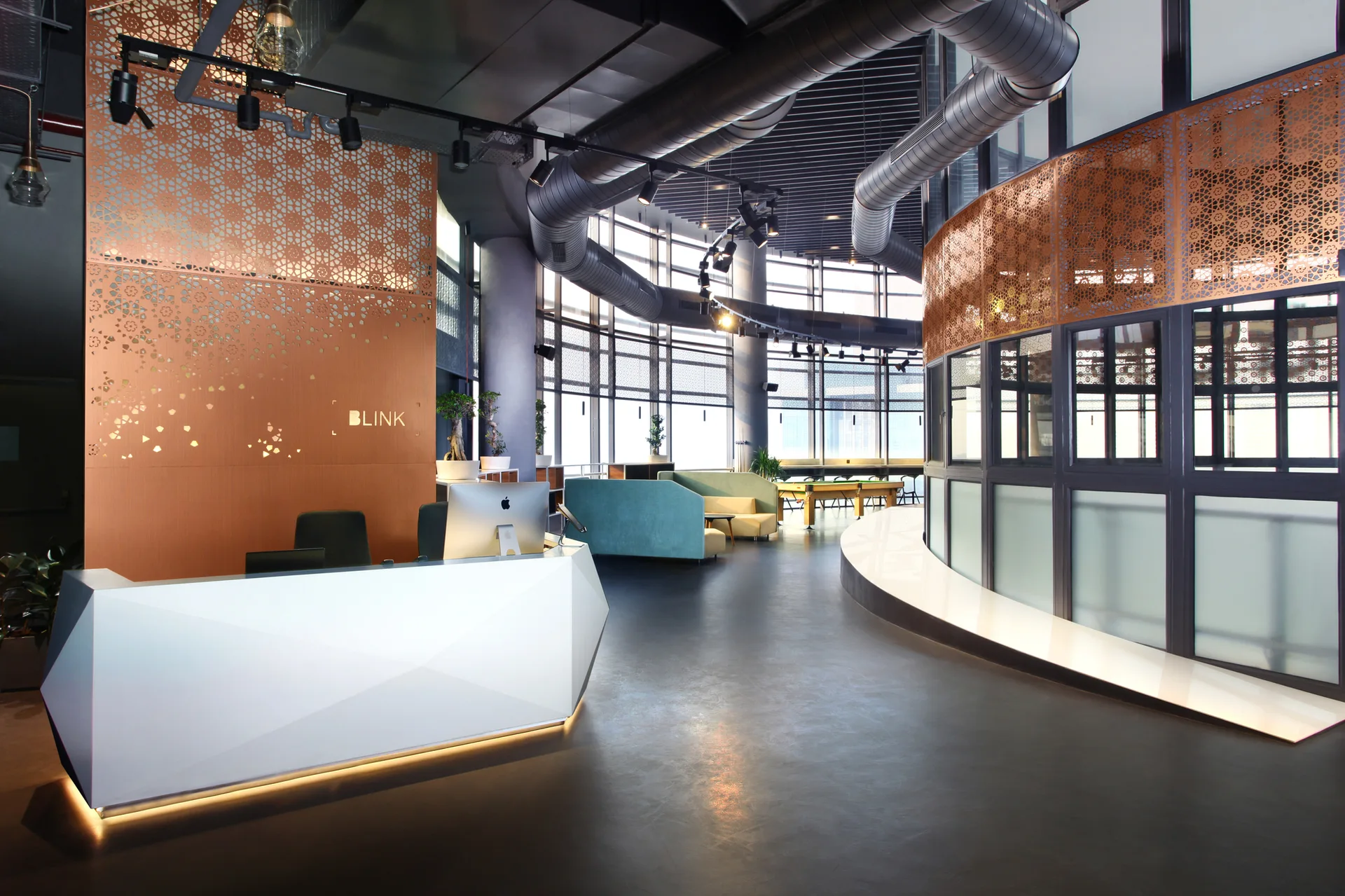 A spacious office lobby features a faceted white reception desk, laser-cut copper screens, and exposed silver ceiling ducts above a curved glass window wall.