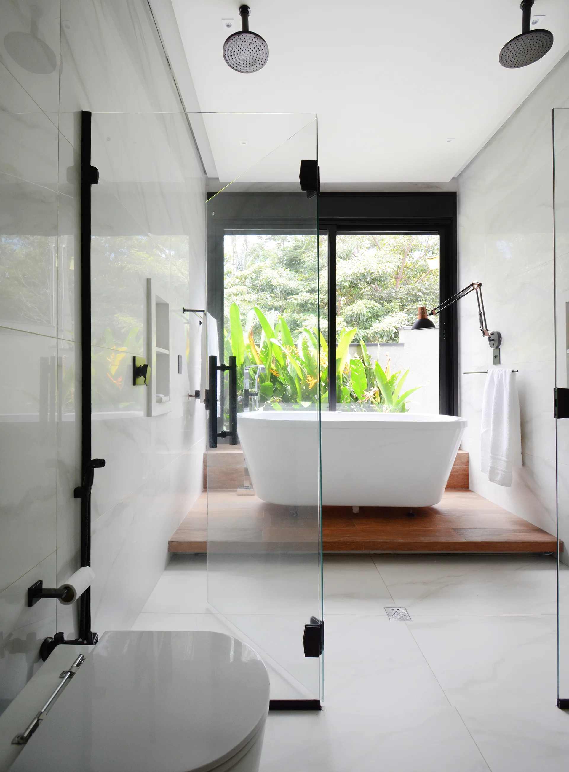 A modern white marble bathroom featuring a freestanding tub on a wooden platform next to a large window overlooking lush green plants.