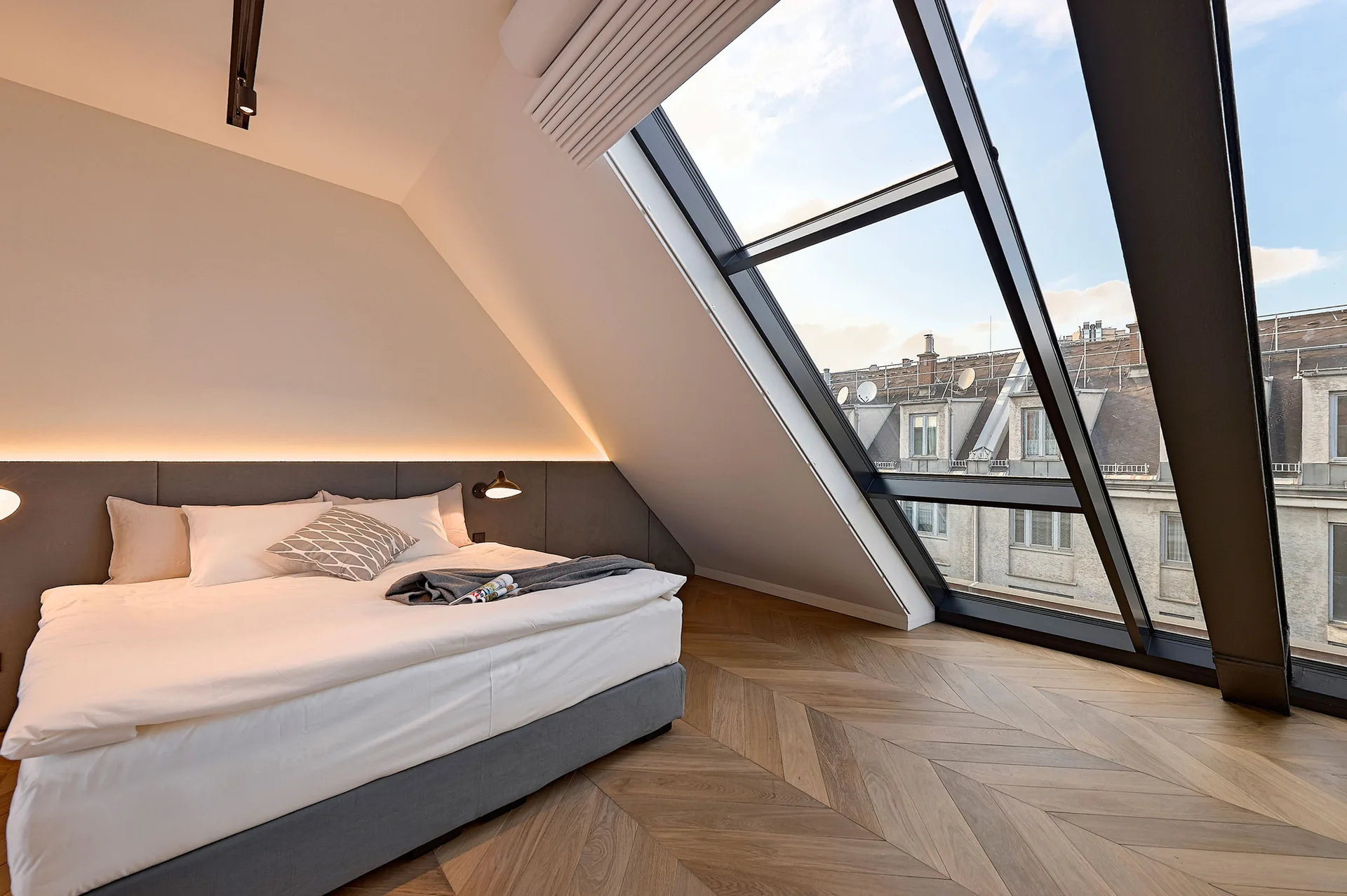 A modern attic bedroom features a low platform bed beneath a sloped ceiling, a large angled skylight window, and herringbone wood flooring.