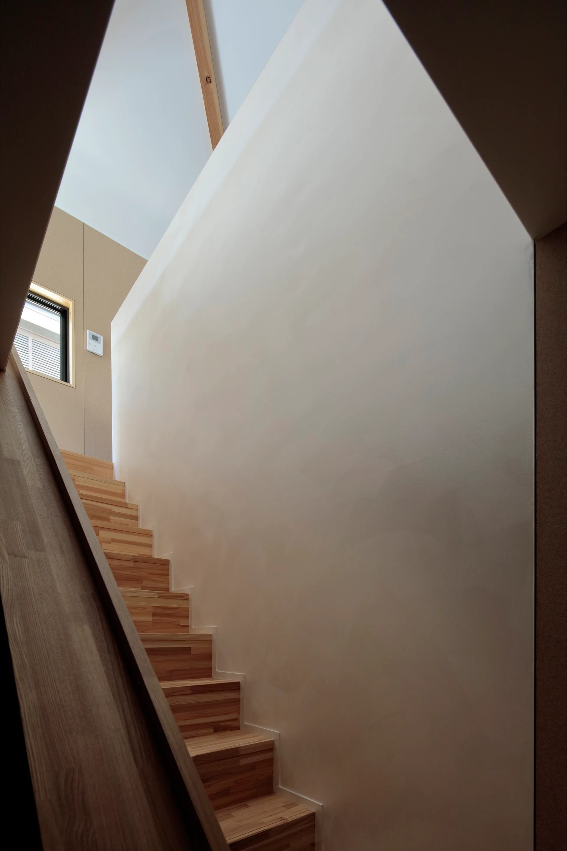 A minimalist wooden staircase with light oak treads climbs upward alongside a tall, smooth white plaster wall under natural light.