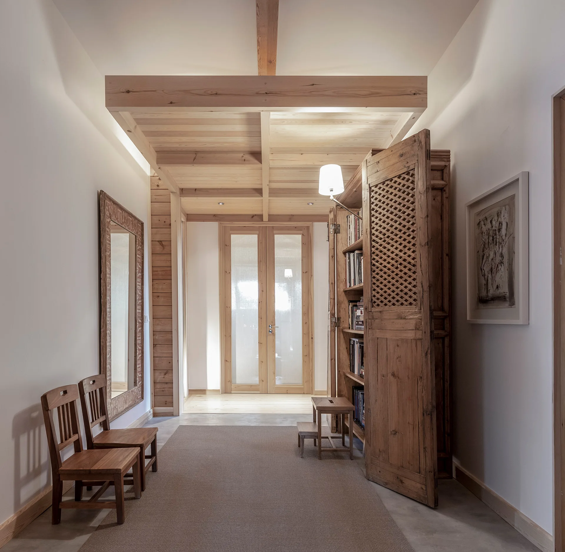 A hallway featuring exposed wood ceiling beams, a large lattice-door cabinet, and two wooden chairs opposite a framed mirror.