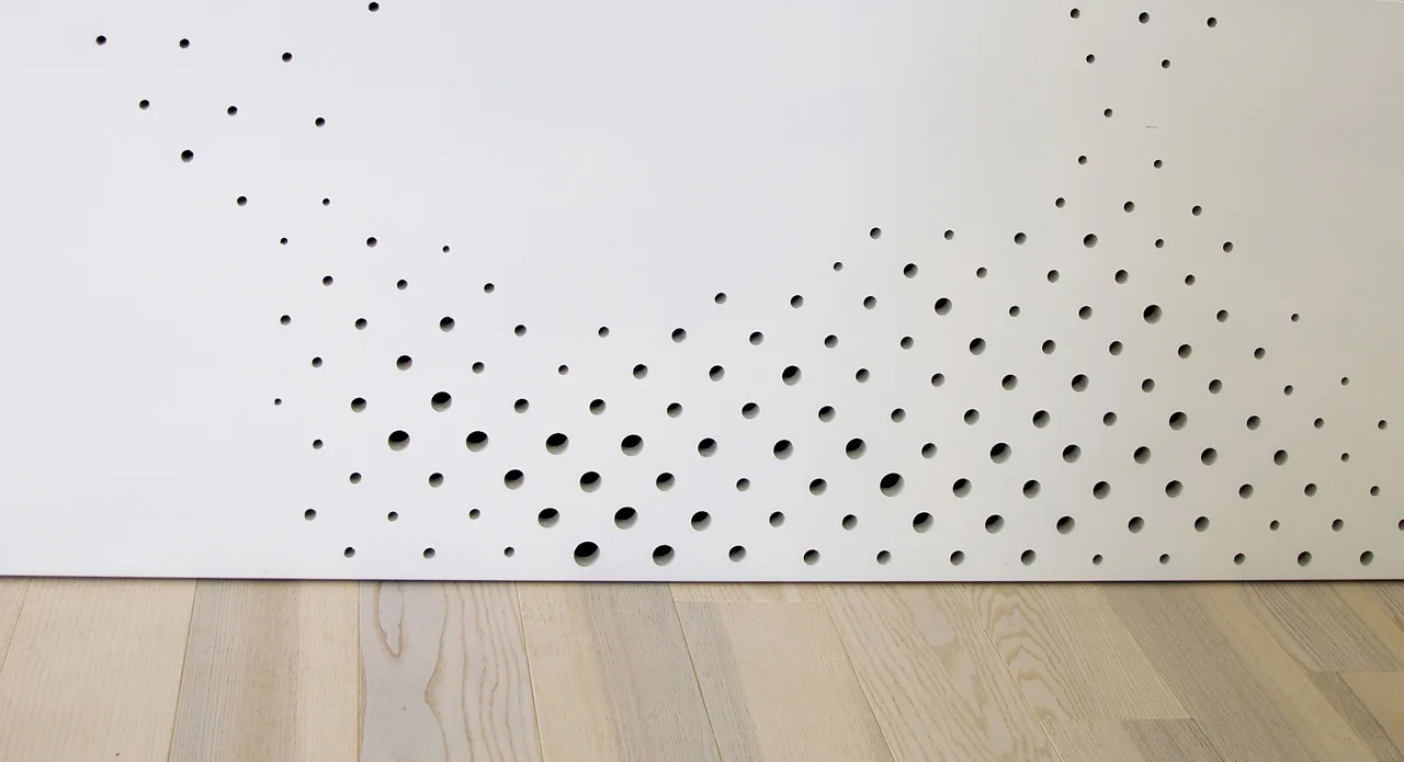 A white perforated panel with varying sizes of circular cutouts sits above light wood plank flooring.