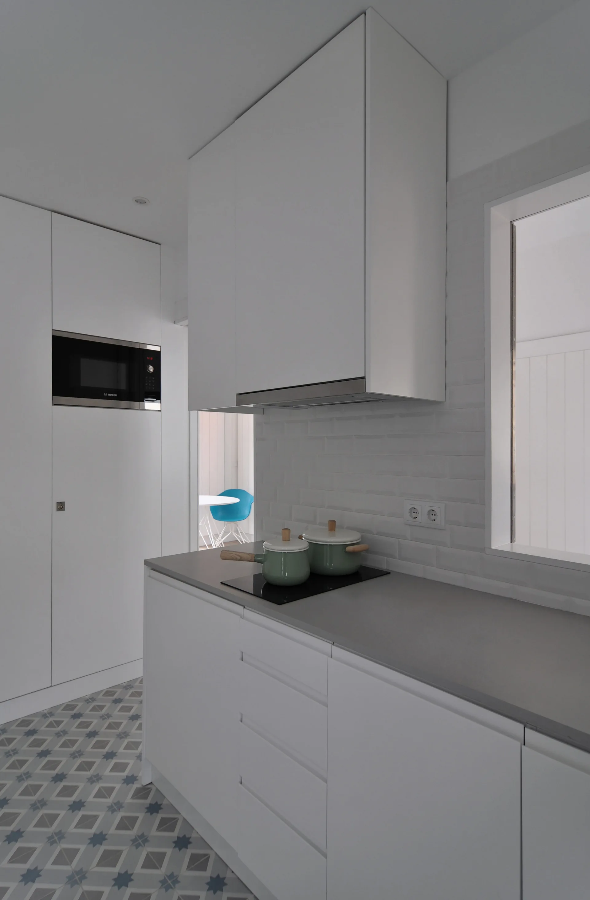 A modern white kitchen features flat-front cabinetry, a gray countertop with a cooktop, and blue-and-gray geometric patterned floor tiles.