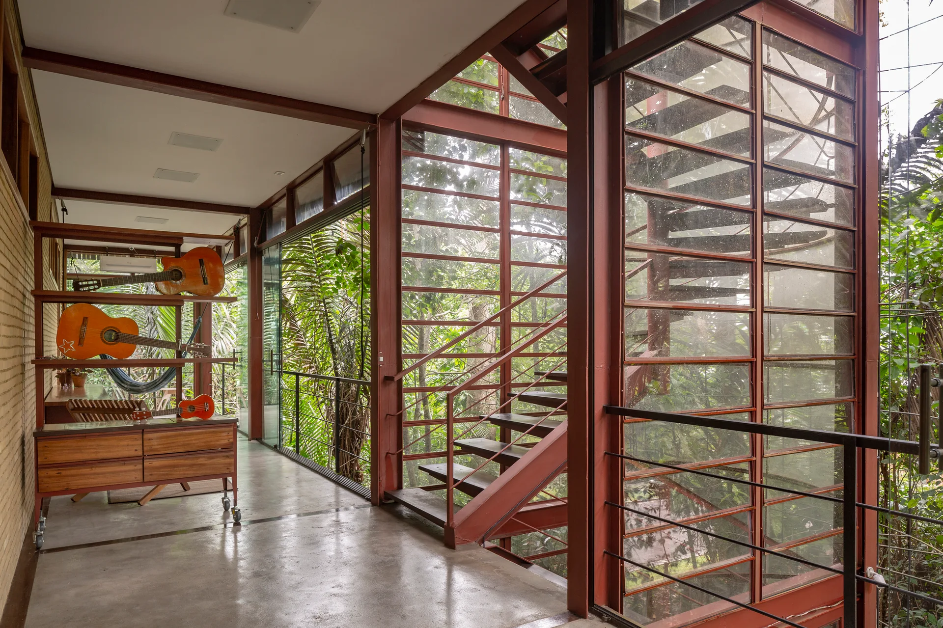 A modern hallway with a polished concrete floor, exposed brick wall, red steel frame windows, and a spiral staircase next to displayed guitars.