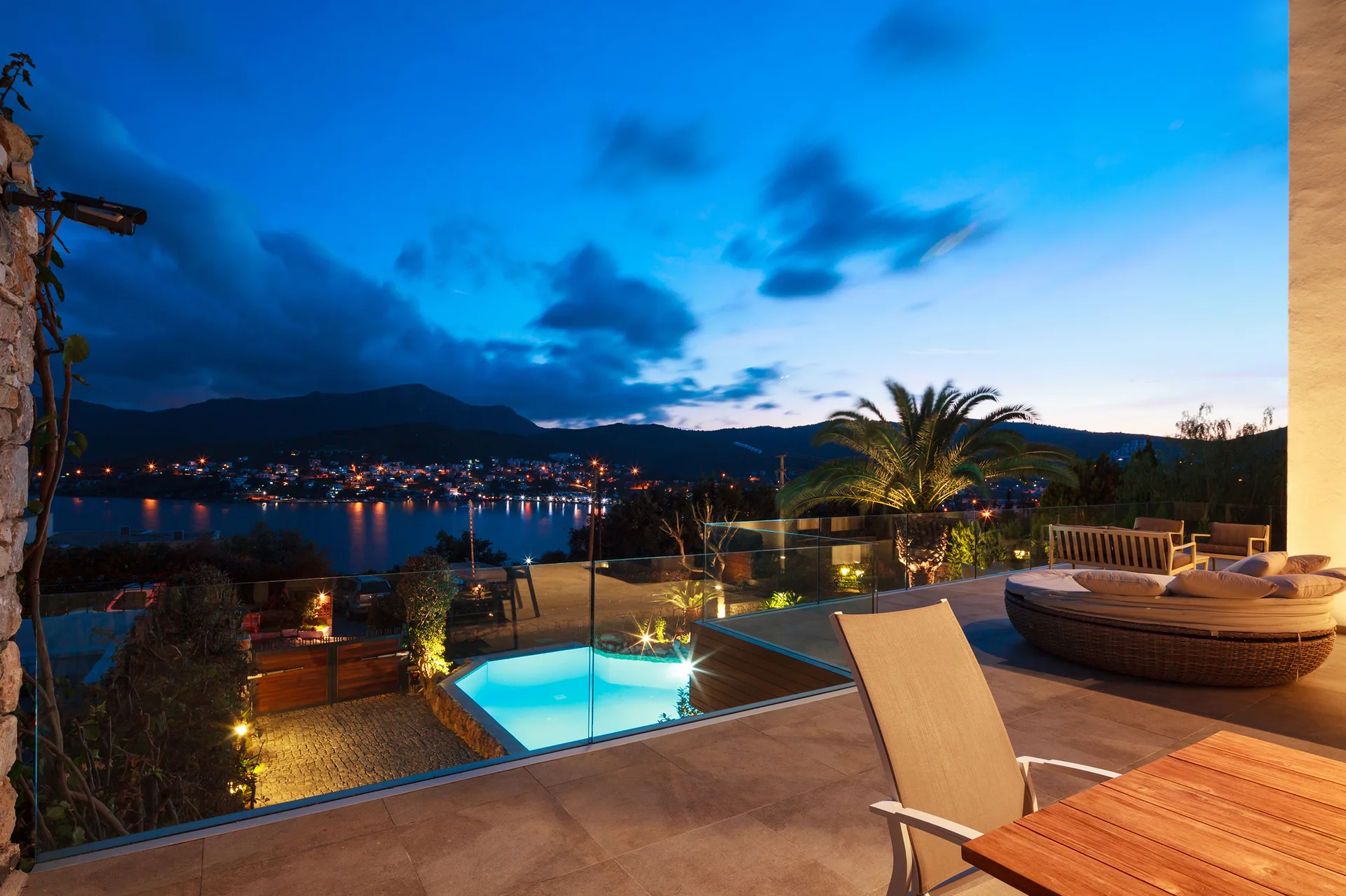 An outdoor stone patio at dusk overlooks a bay with a circular daybed, glass railings, and a lower-level pool.