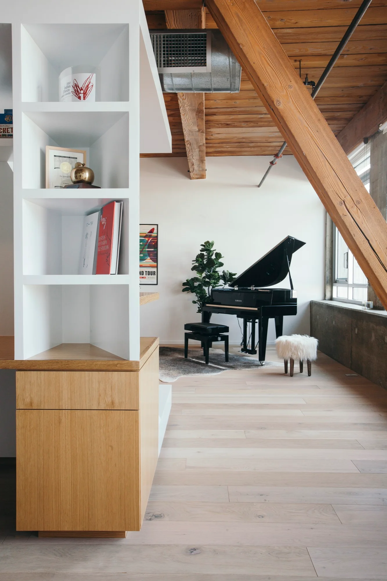 An industrial loft room featuring light wood flooring, exposed ceiling beams, a white built-in shelf, and a black grand piano near a window.