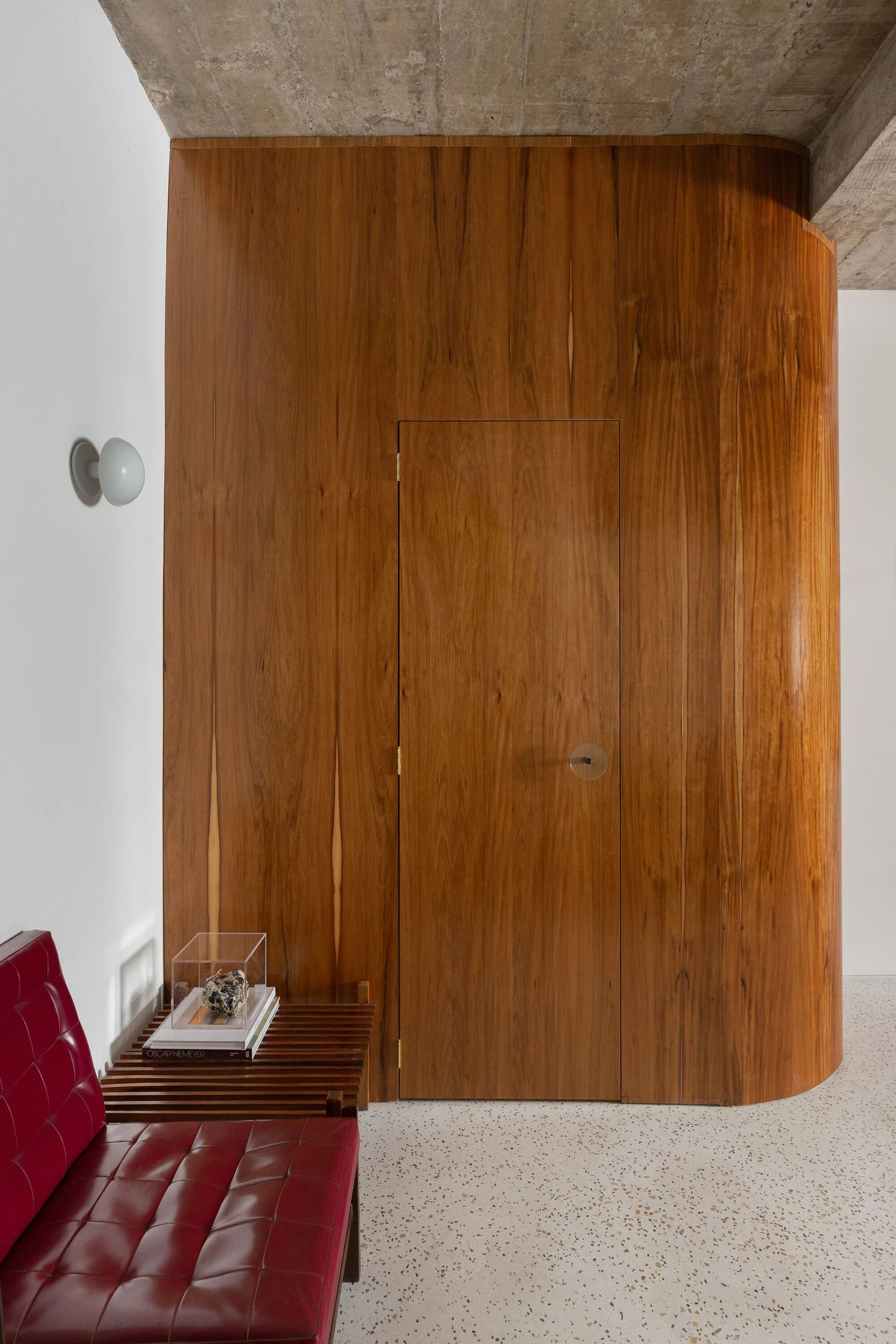 A minimalist entryway features a curved wood-paneled wall with an integrated door, terrazzo flooring, and a red tufted leather seat.