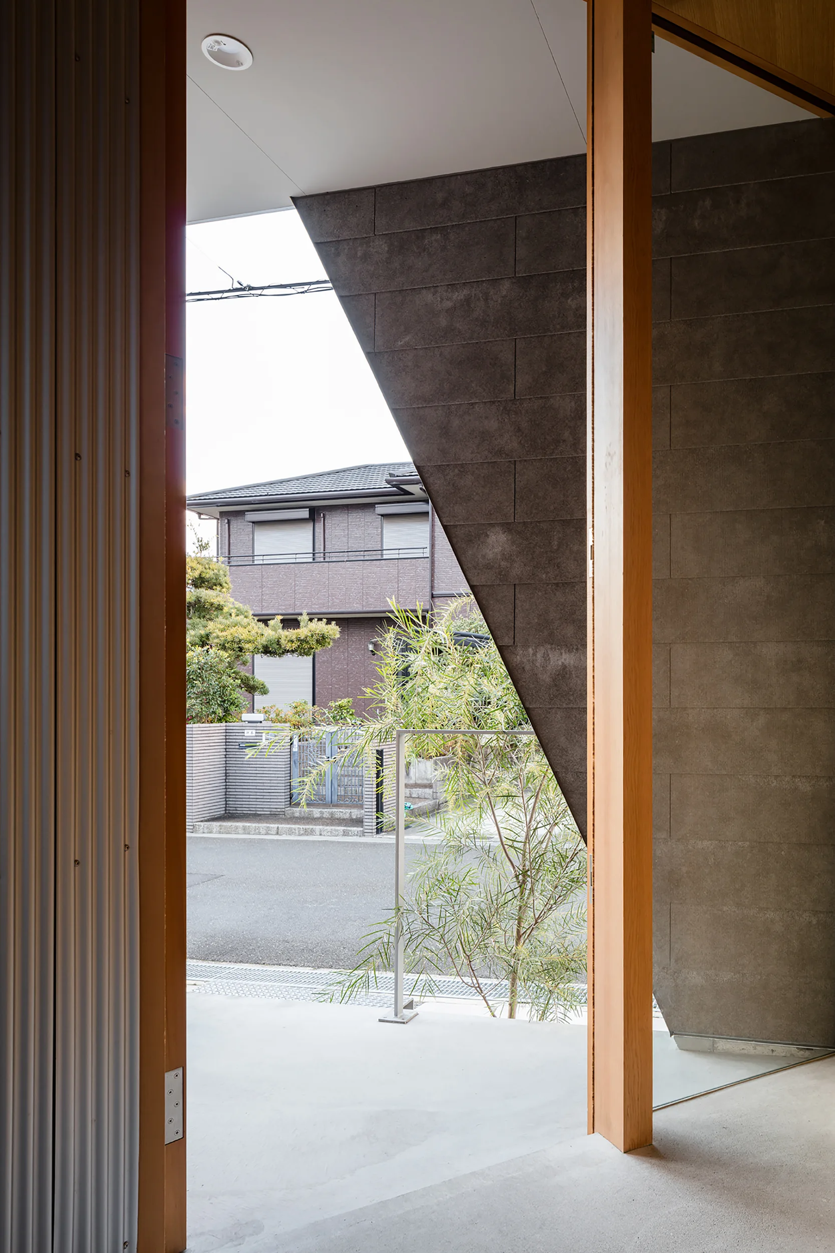 A modern entryway with light wood columns, smooth concrete flooring, and a dark grey tiled wall framing a view of a residential street.