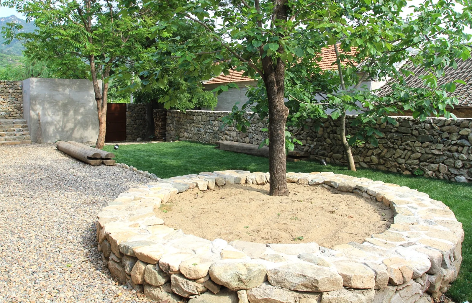 A landscaped garden features a circular stone tree planter filled with sand, a gravel pathway, a green lawn, and a dry-stacked stone retaining wall.