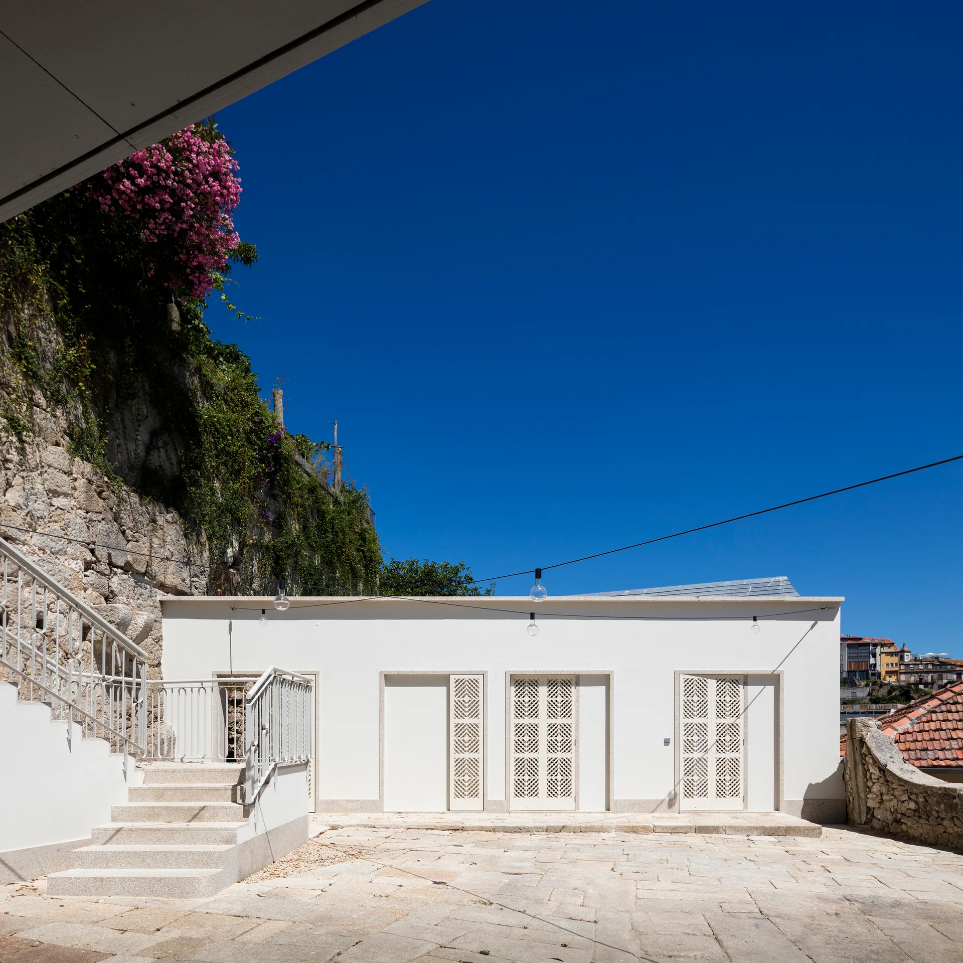 A paved stone courtyard features a white plastered building with three decorative latticed doors next to a staircase and a verdant cliffside.