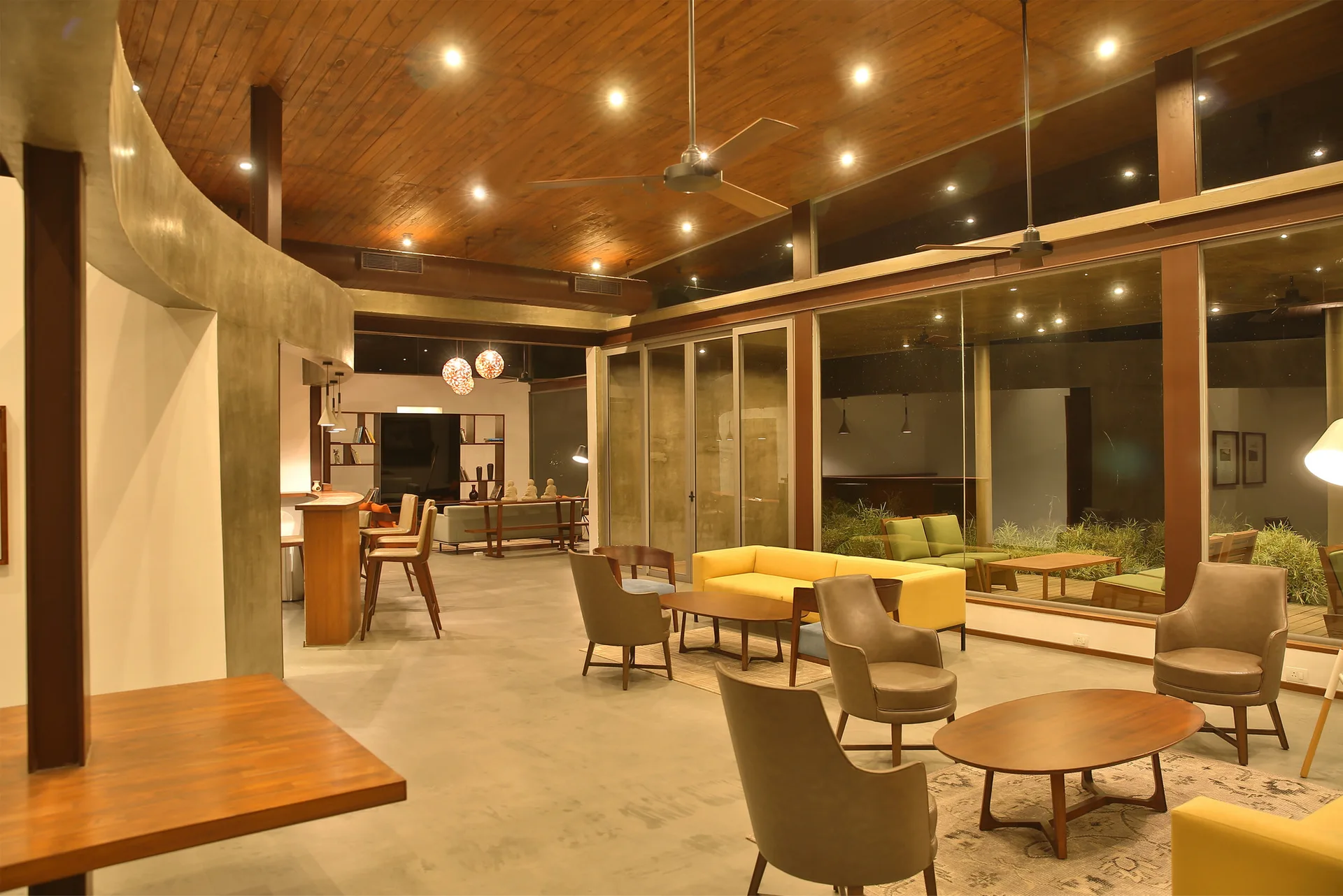 Spacious living room features a vaulted wood-paneled ceiling, concrete floors, yellow and grey seating areas, and floor-to-ceiling windows.