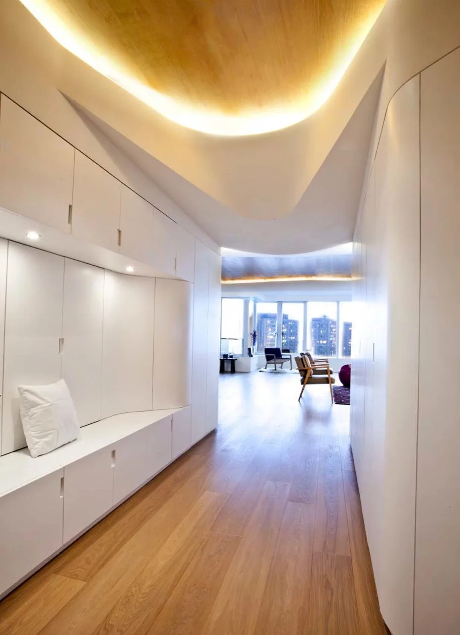 A minimalist white hallway featuring custom built-in cabinetry with an integrated bench, light wood floors, and a curved ceiling with glowing cove lighting.