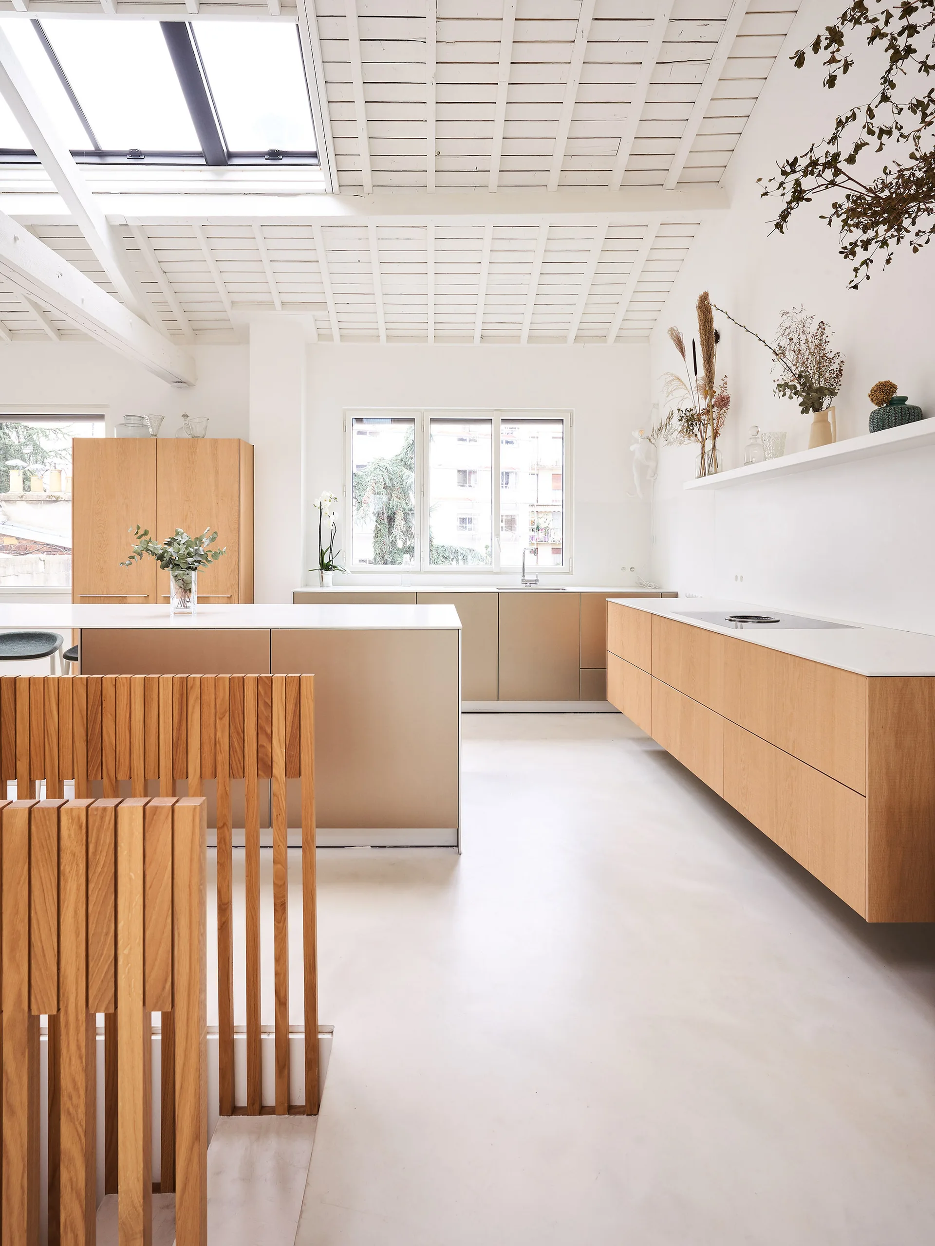 A bright modern kitchen features light wood cabinets, a white island, and a slatted wood partition under a white beamed ceiling with a skylight.