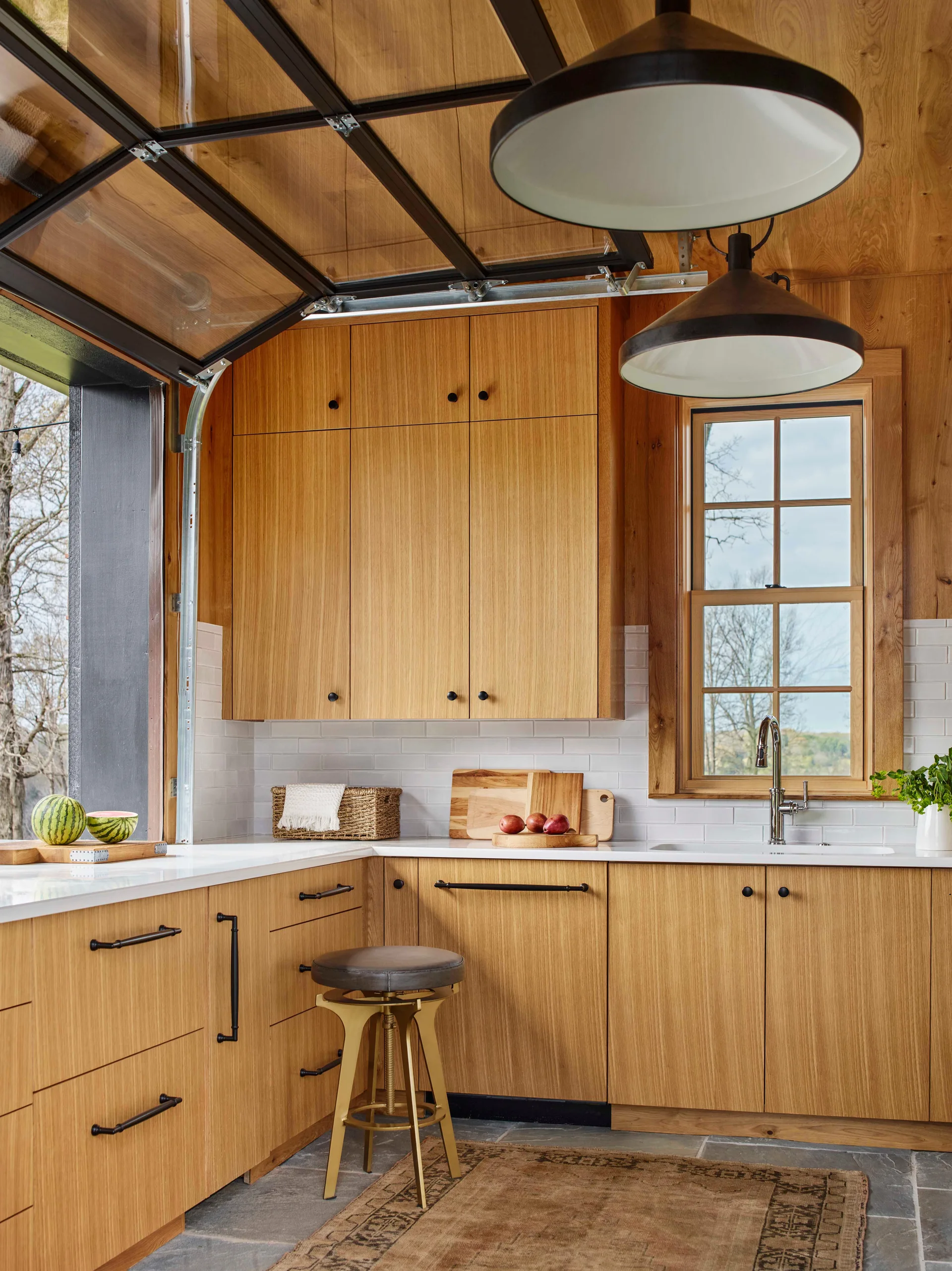 A kitchen with natural wood cabinetry, white countertops, a grey stone tile floor, and an open glass garage-style window looking outside.