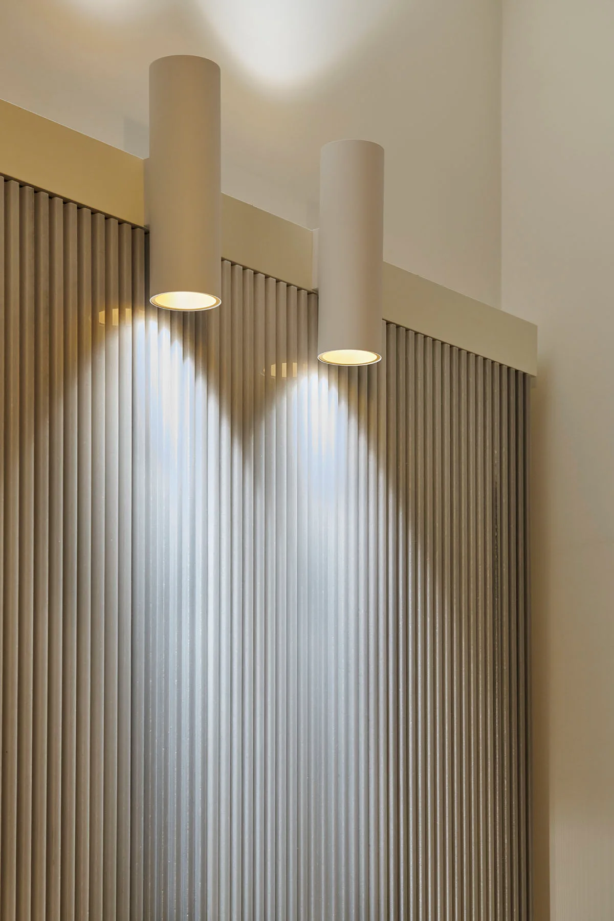 Two matte beige cylindrical downlights cast warm light down the vertical ridges of a fluted wall panel.