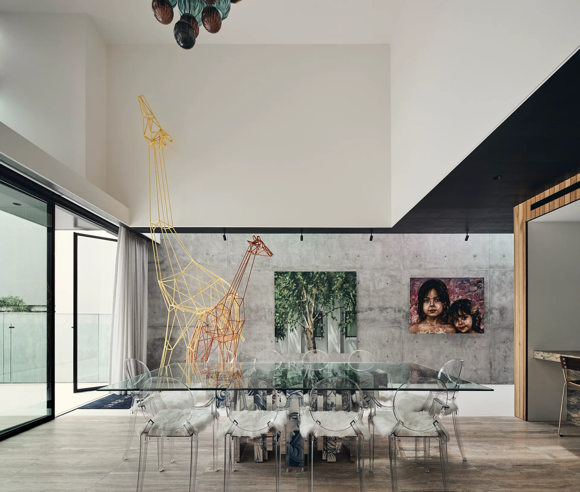 A contemporary dining area features a glass-topped table, transparent acrylic chairs, and a large wire giraffe sculpture against a concrete wall.