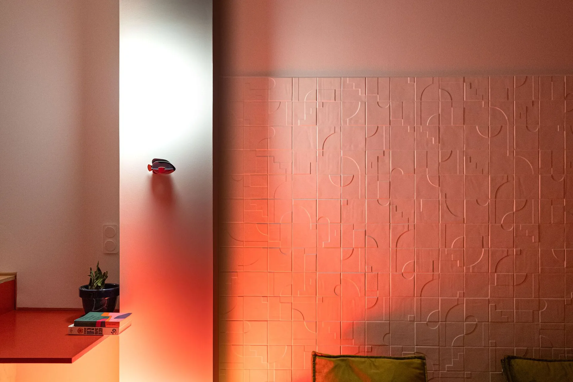 Matte pink wall panels with a raised geometric pattern are illuminated by a warm orange light next to a smooth wall with a red shelf.
