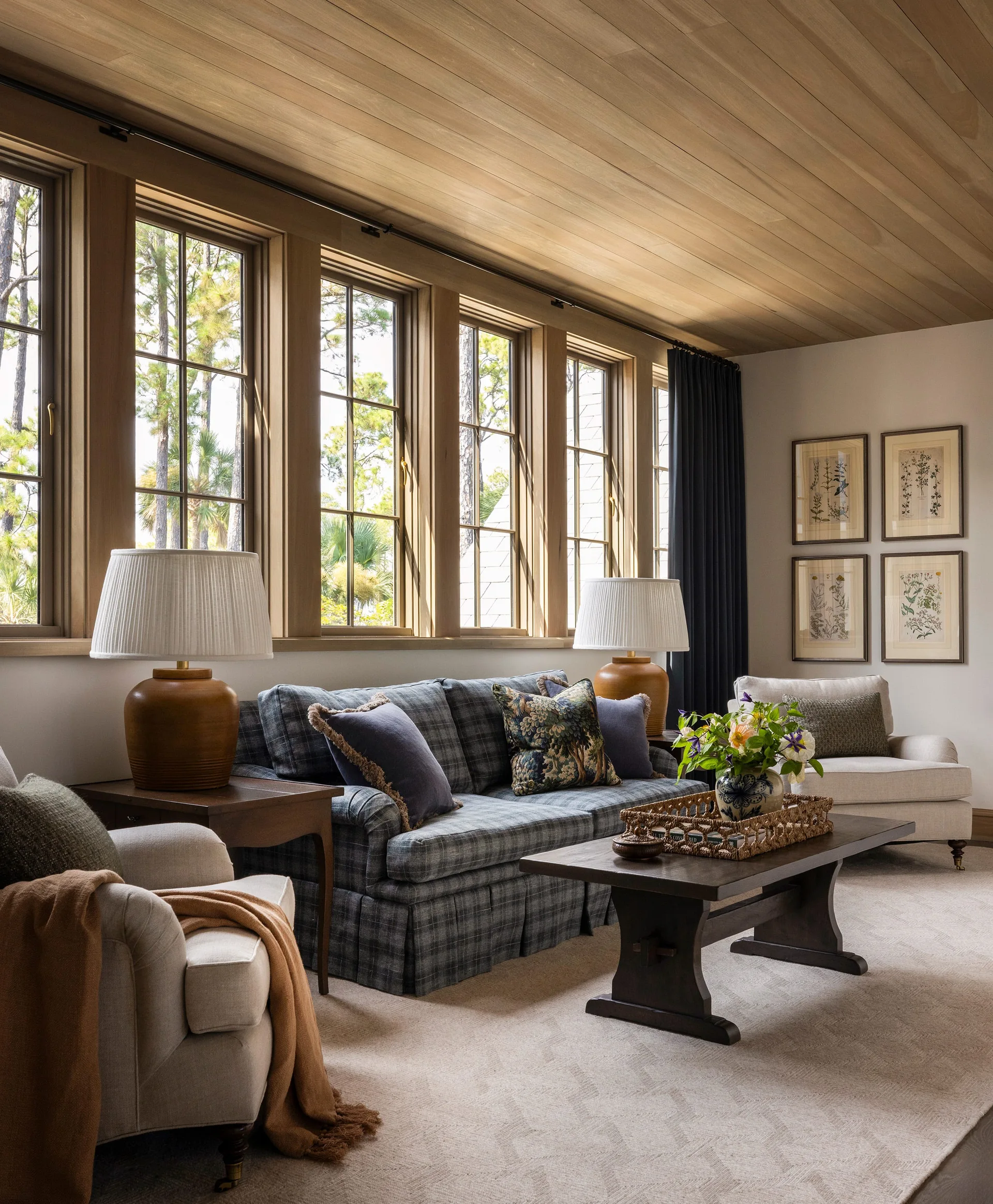 A comfortable living room features a plaid sofa, a dark wood coffee table, and large windows beneath a warm wood plank ceiling.