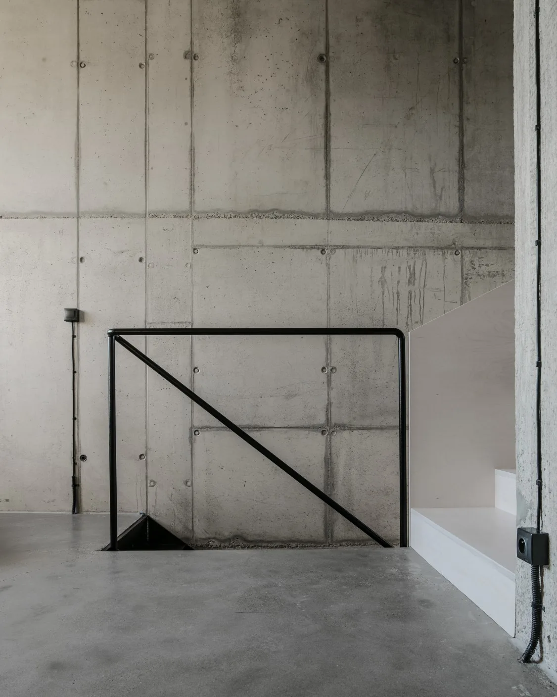 A raw concrete wall serves as the backdrop for a simple black metal handrail next to light-colored wooden stair steps on a concrete floor.