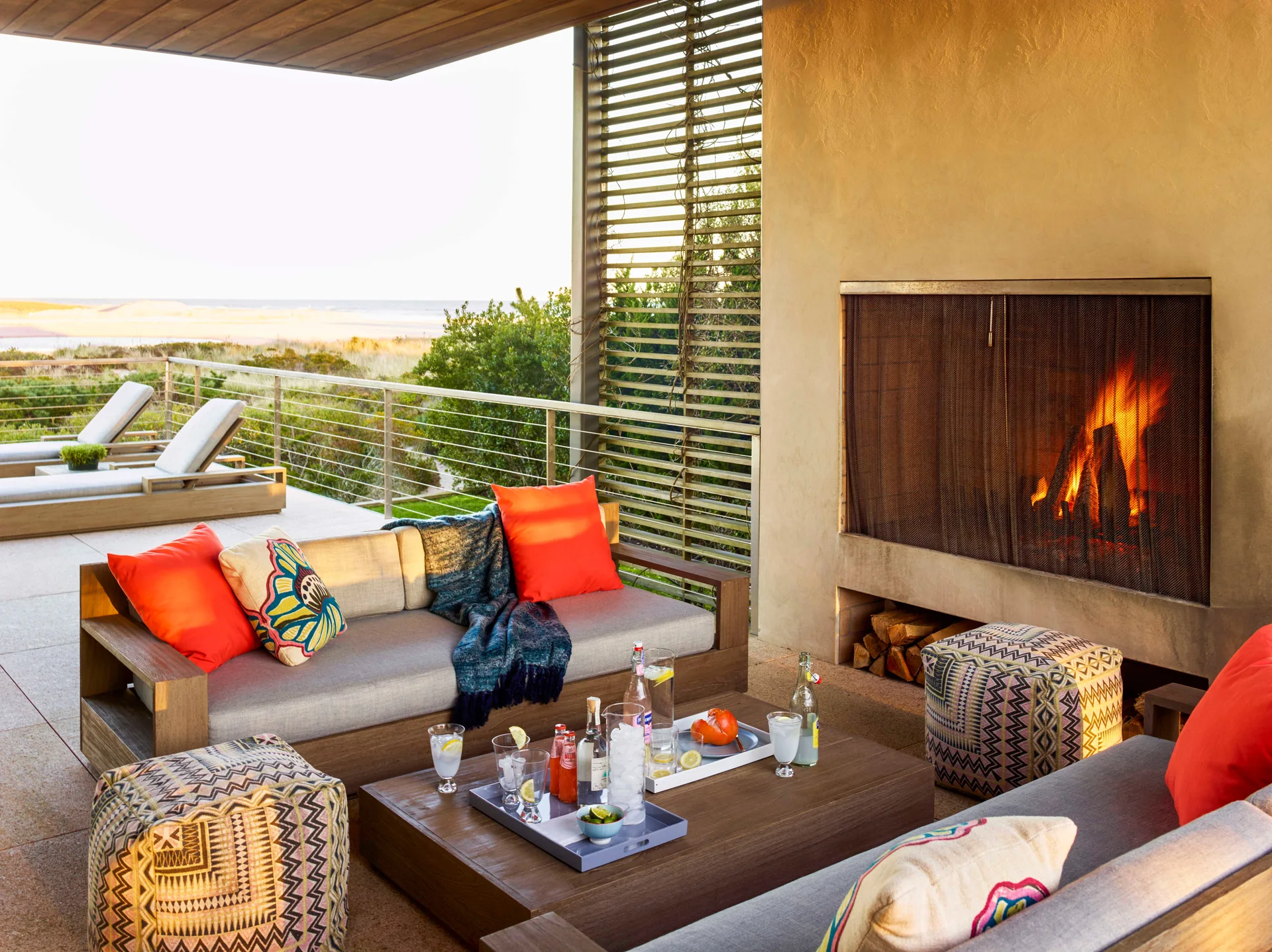 A covered outdoor terrace features wood frame sofas with orange pillows, a stone fireplace with a lit fire, and a view of the ocean.