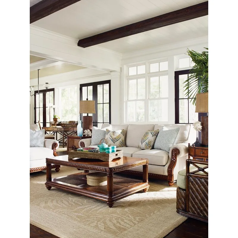 A coastal living room features a cream sofa with carved wood details, a dark wood coffee table, and dark ceiling beams against a white ceiling.