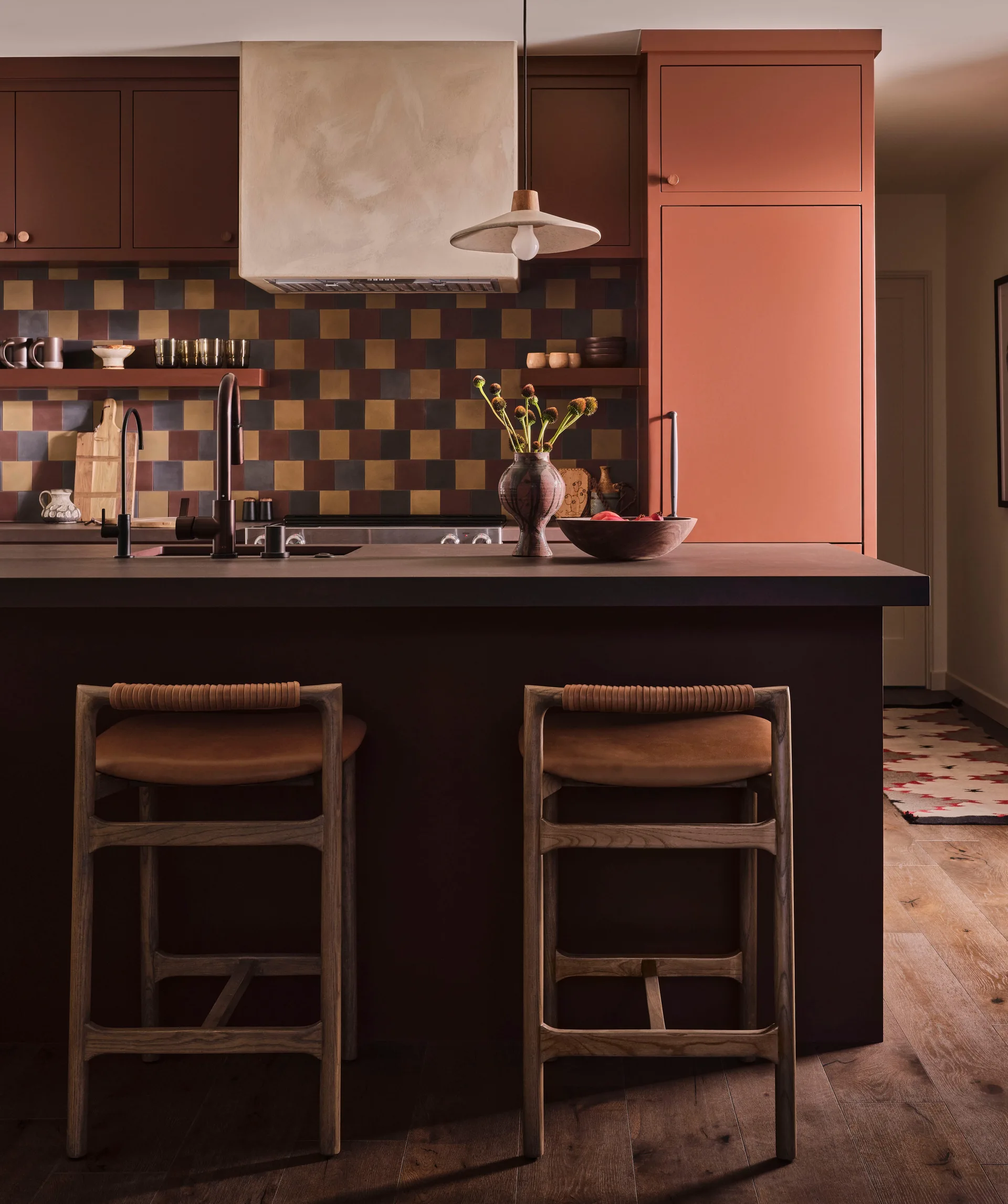 A modern kitchen features a dark island with two wooden bar stools, warm terracotta cabinets, and a multi-toned checkered tile backsplash.