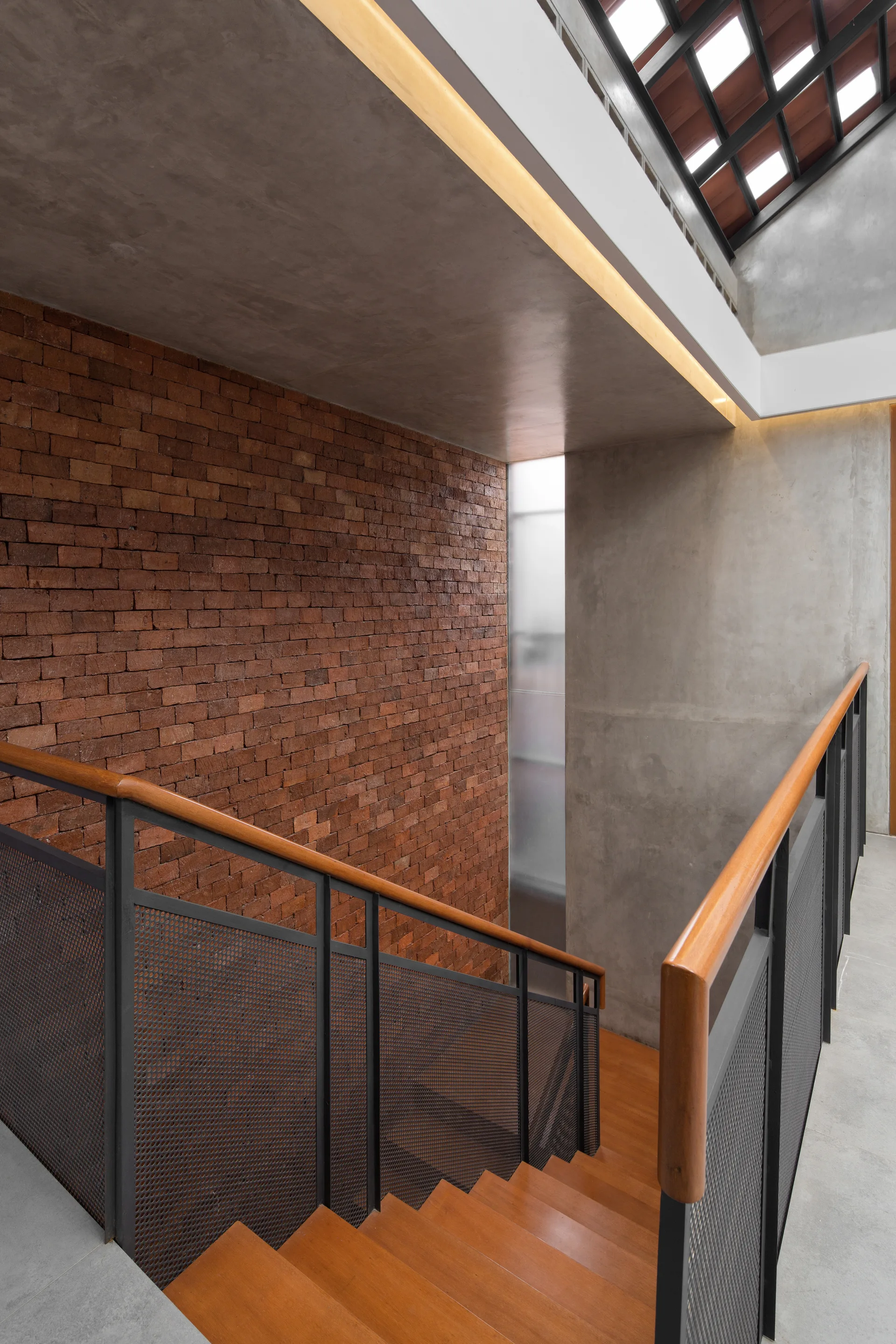 A wooden staircase with perforated metal mesh railings sits between an exposed brick wall and a smooth concrete wall under a skylight.