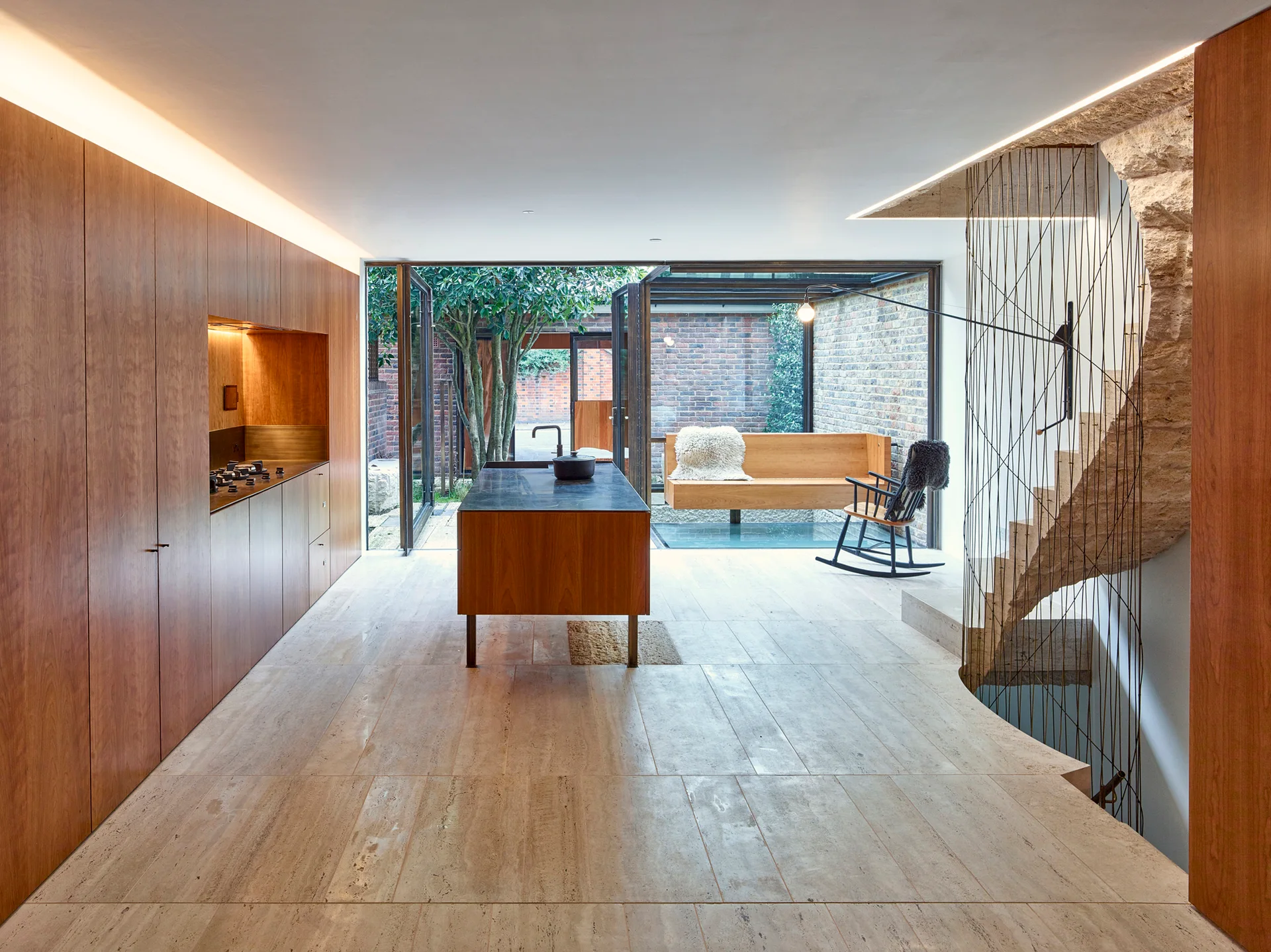 A modern open-plan kitchen with full-height wood cabinetry, a central stone-topped island, travertine tile flooring, and a stone staircase.