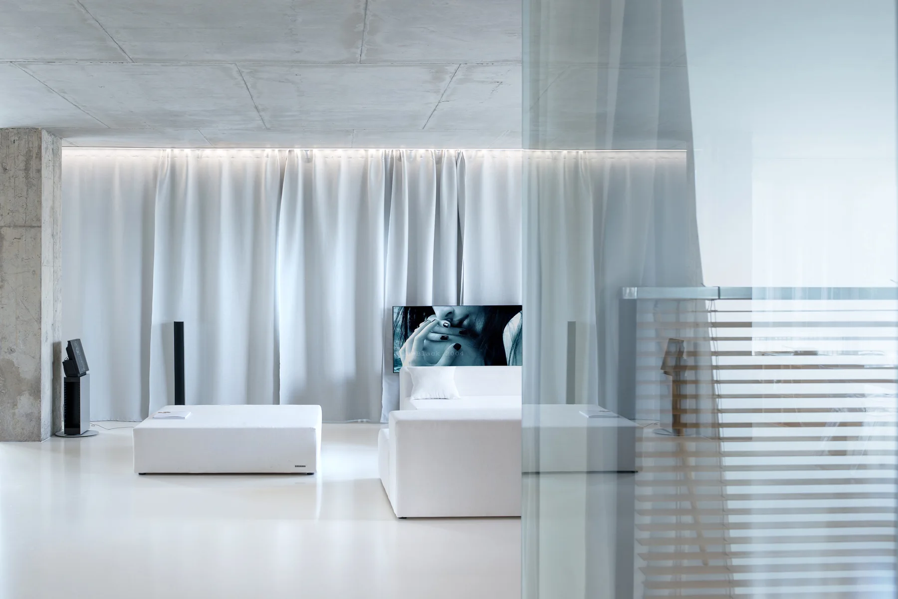 A minimalist living space with a white sofa and ottoman, raw concrete ceiling, and a wall of full-length white drapery.