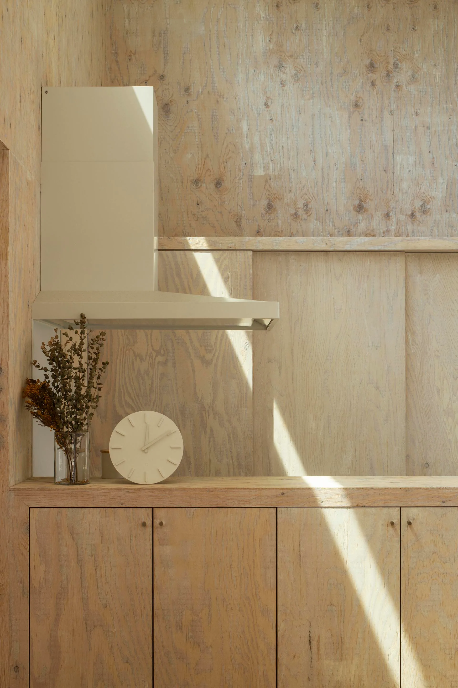 Light plywood wall paneling covers a minimalist kitchen corner with matching wooden cabinetry, an off-white range hood, and a simple cream-colored clock.
