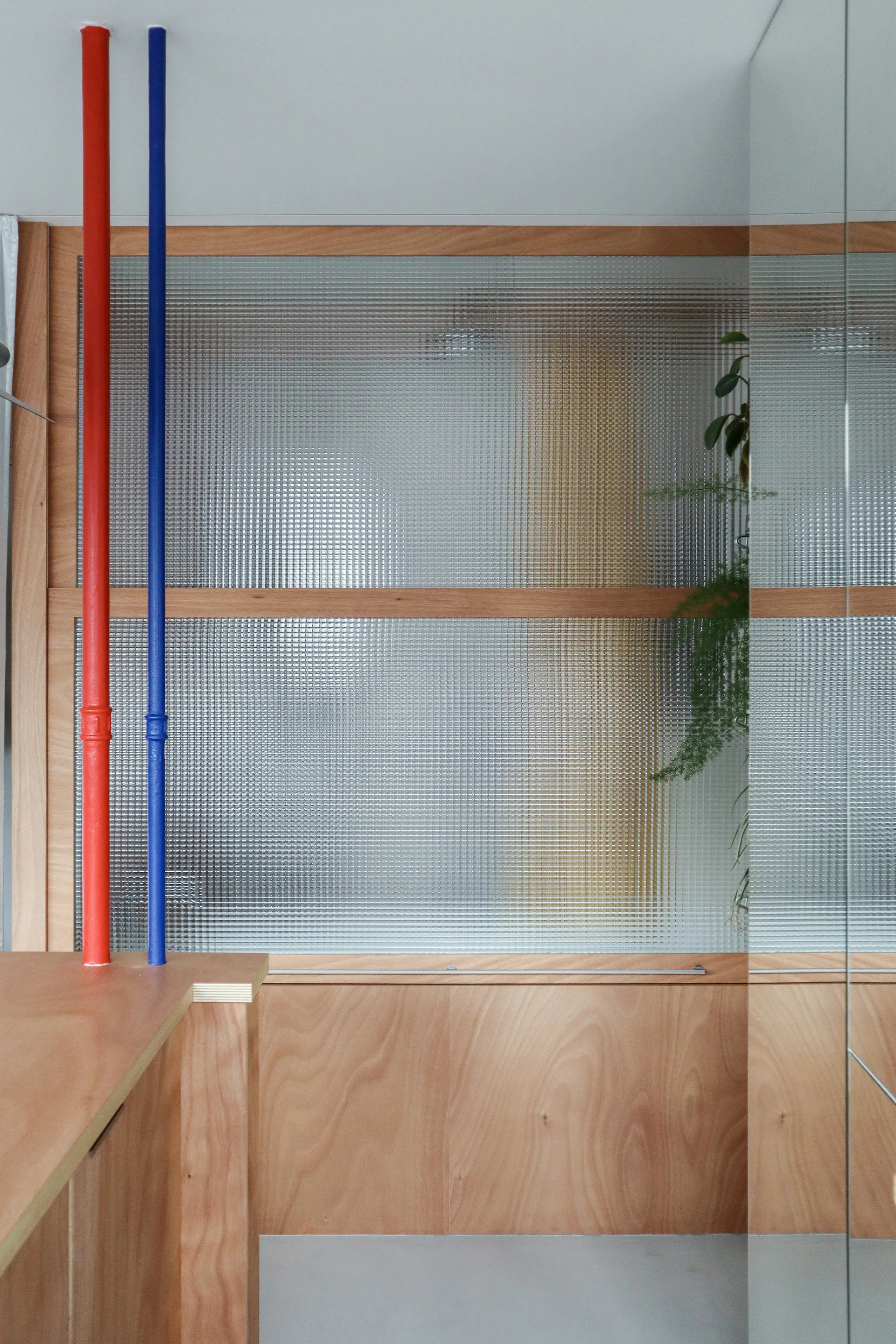 A modern plywood partition featuring a grid-patterned glass screen next to two vertical pipes painted in bright red and blue.
