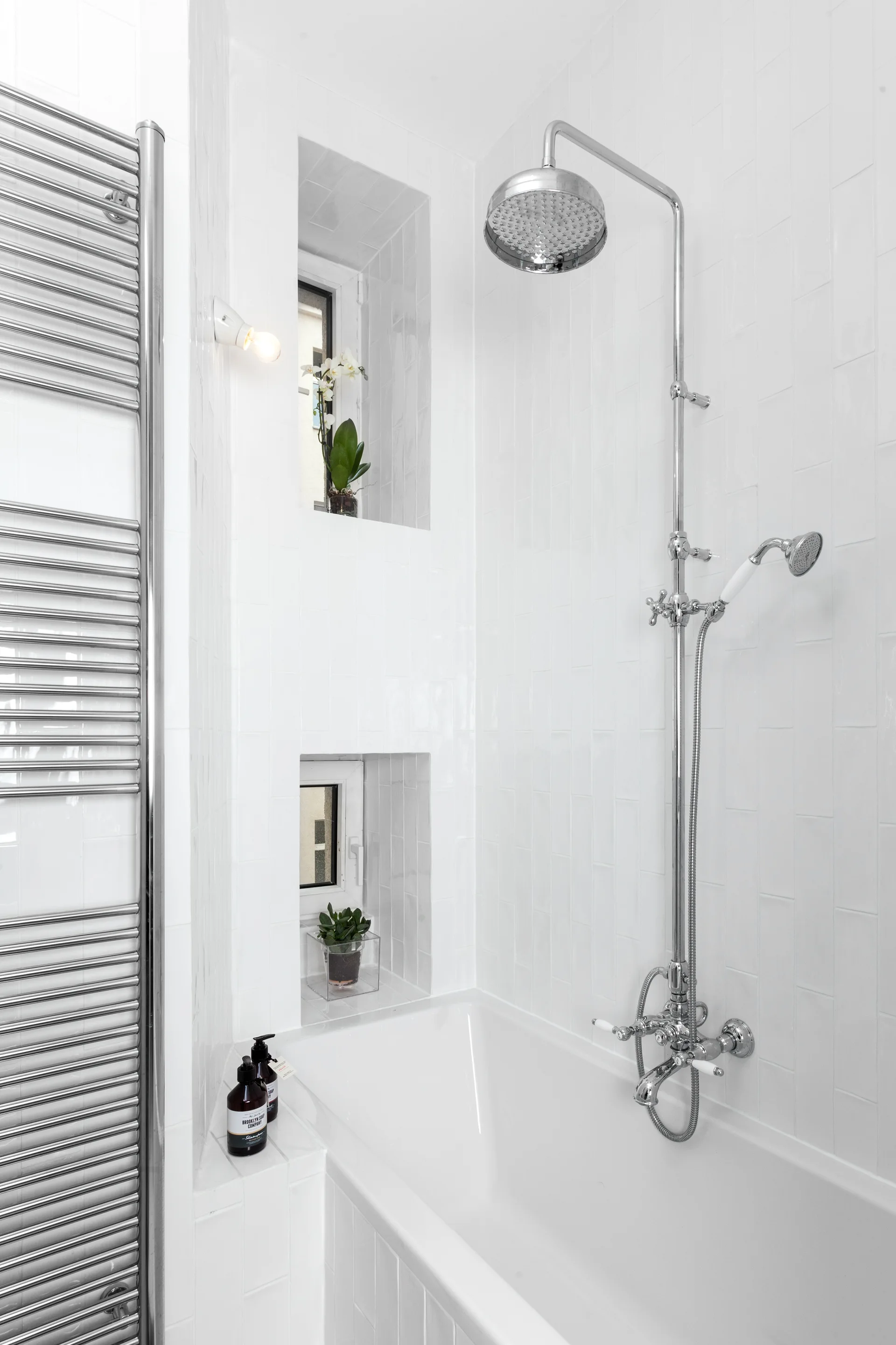 A white tiled bathroom with a chrome shower fixture, a ladder towel warmer, and two stacked recessed window niches with potted plants.