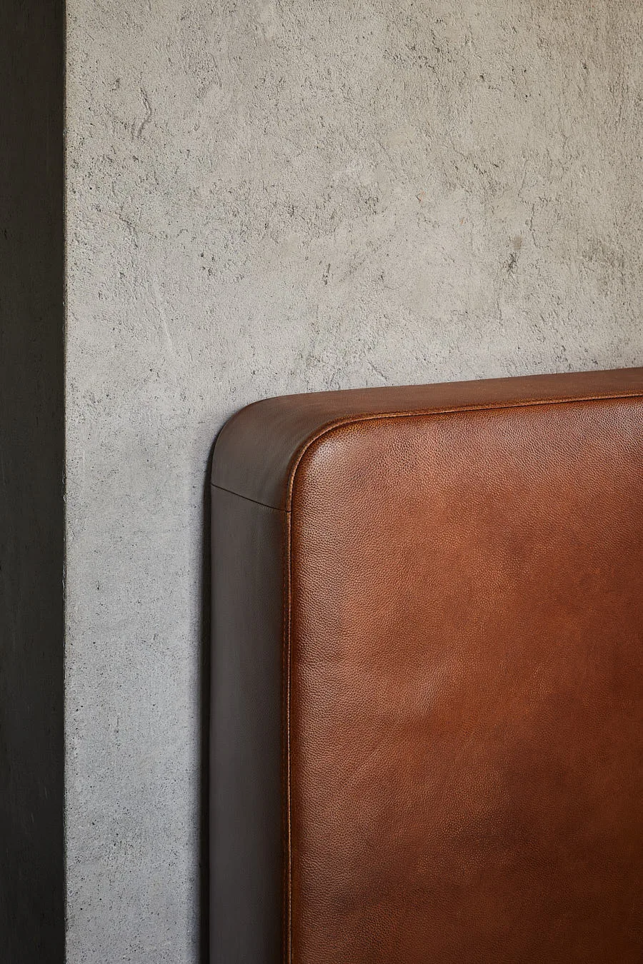 A close-up of a brown leather headboard with a rounded corner positioned against a rough grey concrete wall.
