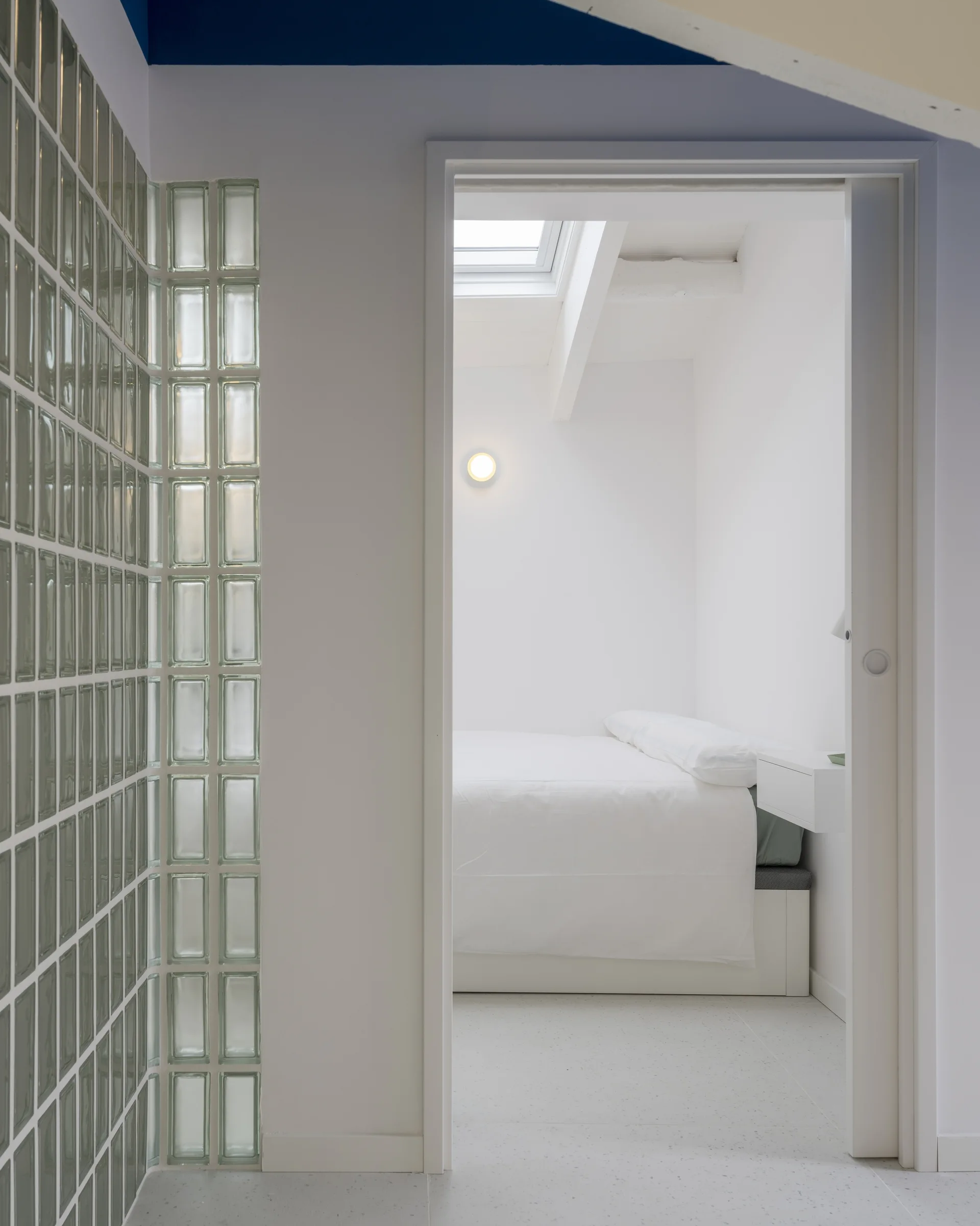 A view through an open doorway into a minimalist white bedroom featuring a skylight and a pale green glass block wall in the foreground.