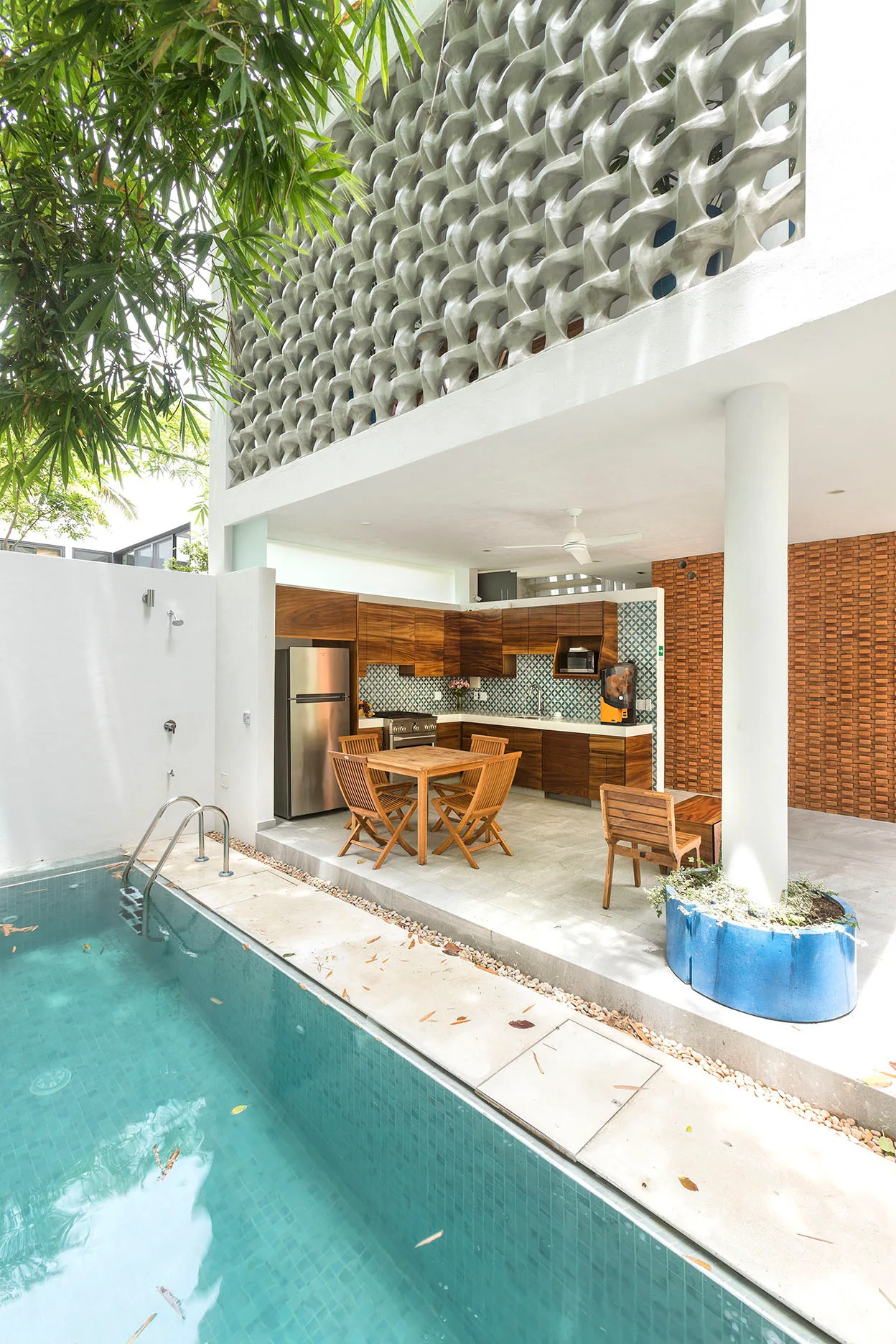 An outdoor kitchen with wooden cabinetry sits next to a swimming pool, featuring a wooden dining set and a decorative concrete screen above.