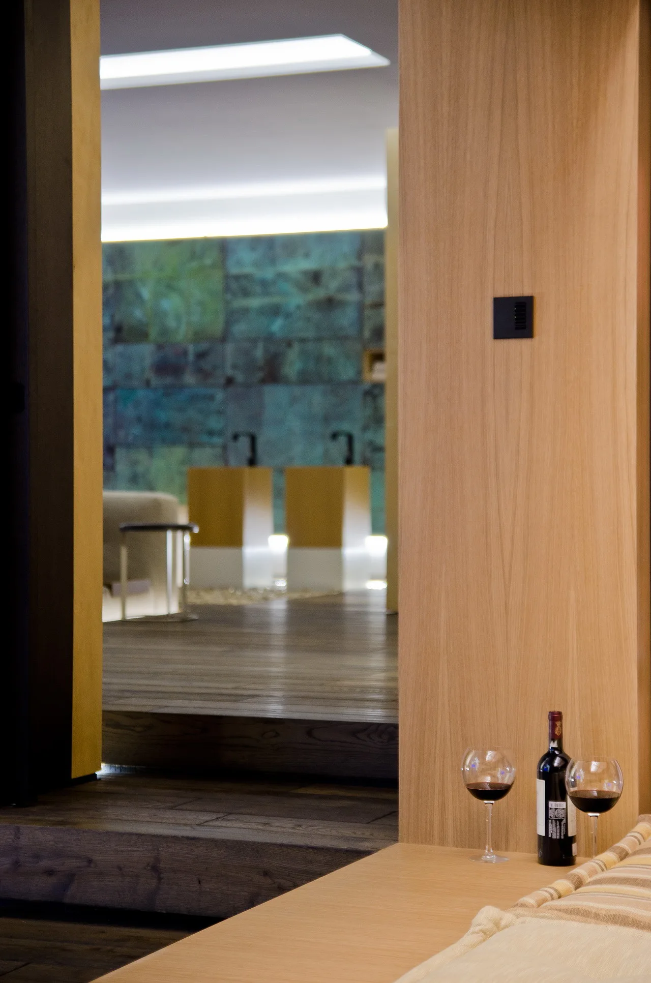 A view through a light wood doorway reveals dark wood steps, a teal textured accent wall, and a foreground with a wine bottle and glasses.