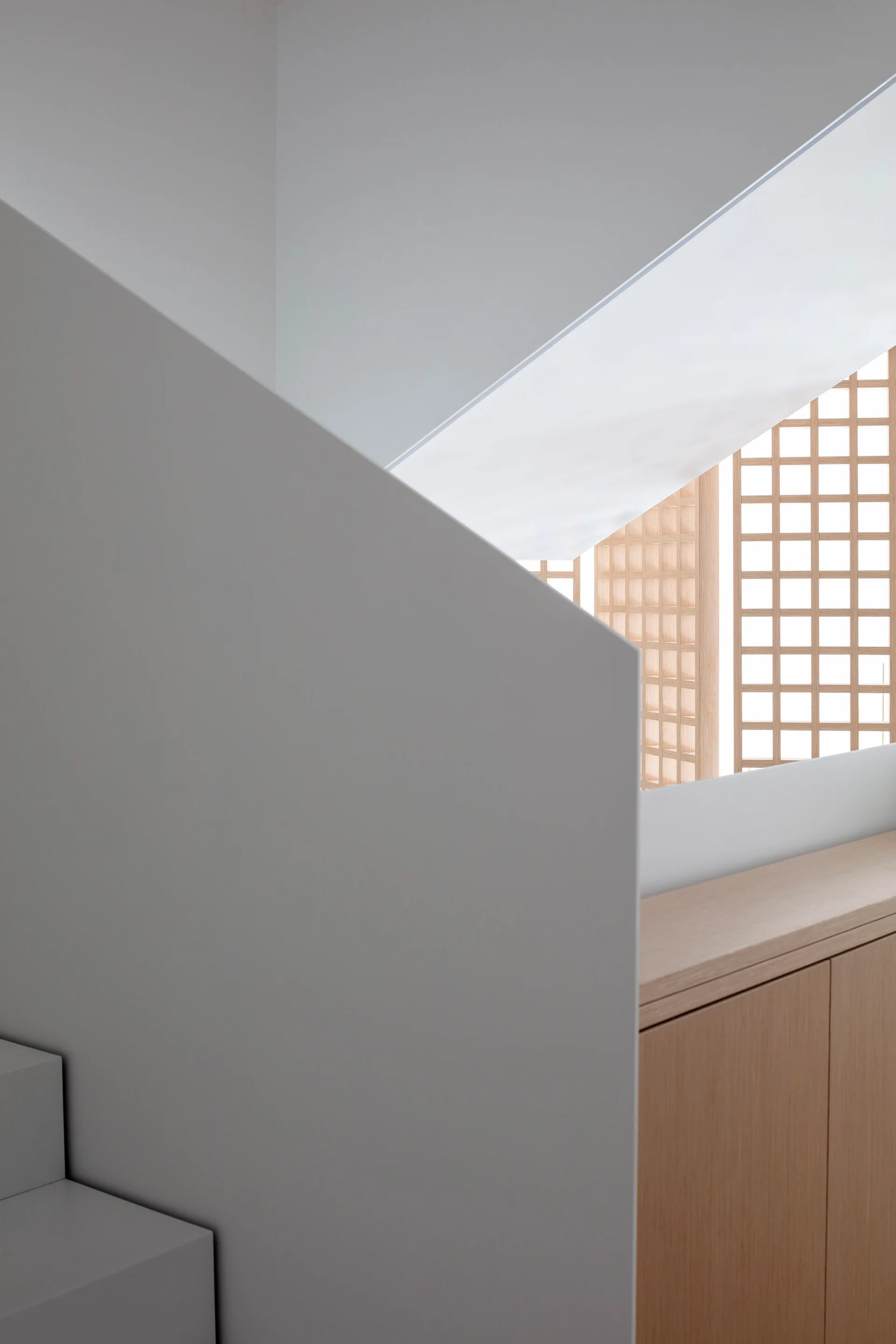 A minimalist interior featuring a white angled staircase wall next to a light wood cabinet and a wooden lattice screen filtering natural light.