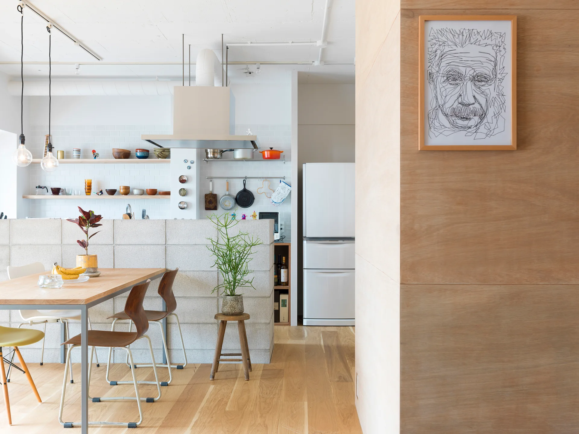 A wooden dining table sits next to a low concrete block partition wall in a bright kitchen with open shelving and white subway tile.