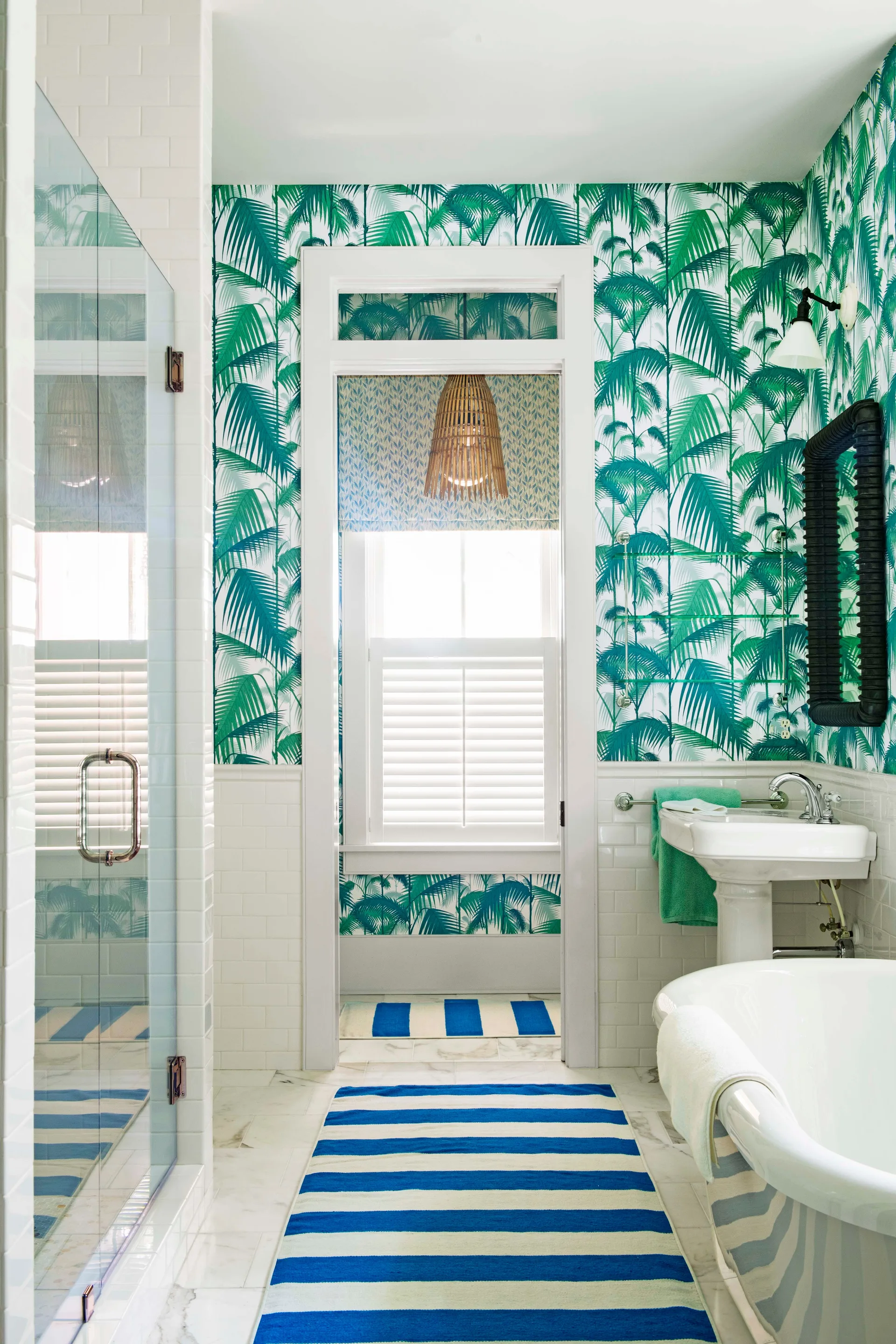 A bathroom featuring green tropical palm wallpaper above white subway tile wainscoting, with a pedestal sink, clawfoot tub, and blue striped rugs.