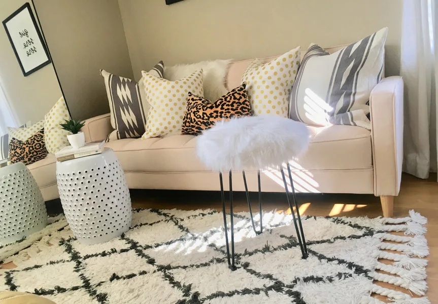A blush pink sofa sits on a shaggy geometric rug, styled with patterned throw pillows and accompanied by a white faux fur hairpin stool.