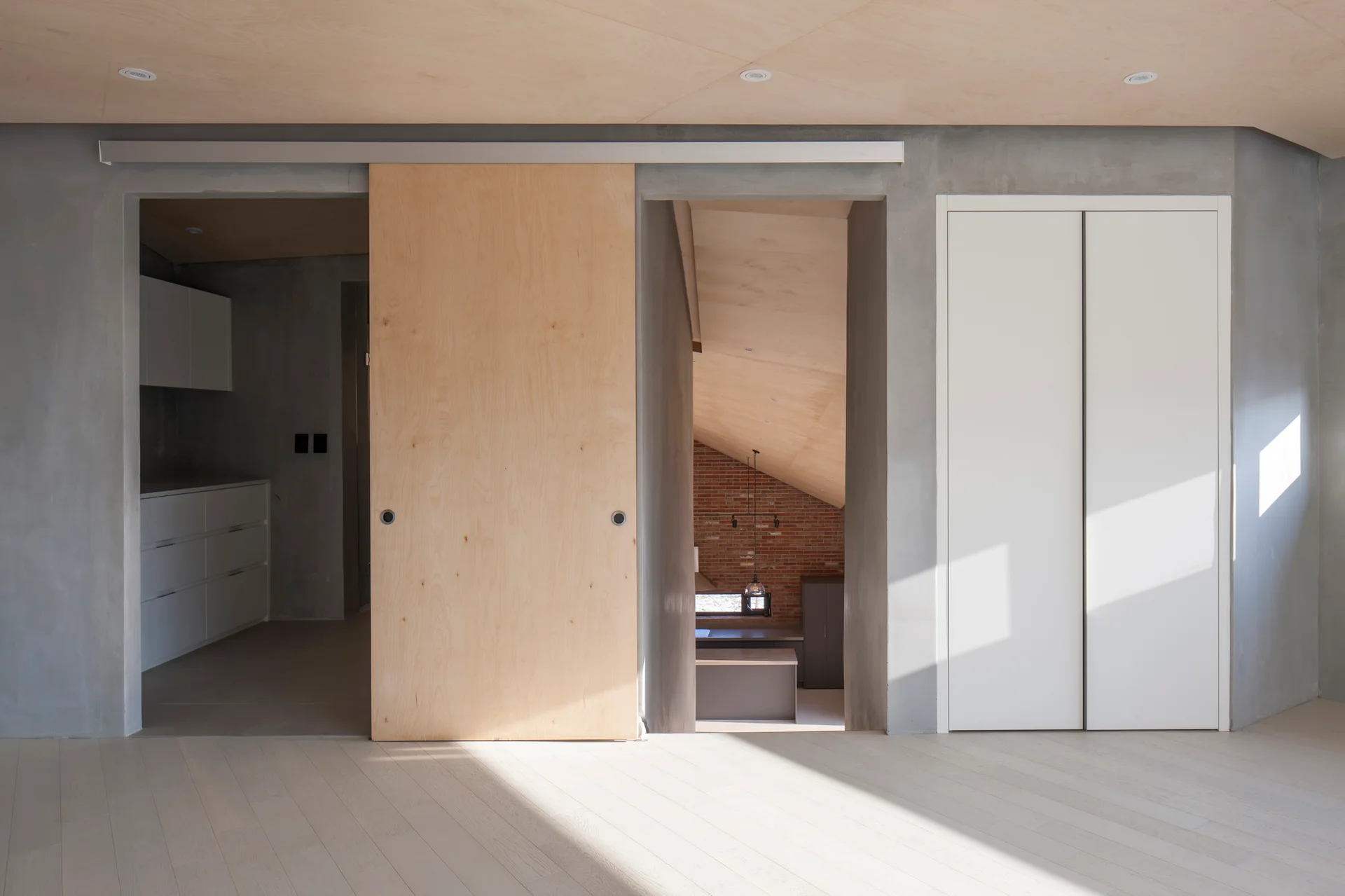 A minimalist room featuring textured grey concrete walls, a light plywood ceiling, and a large wooden sliding door between two door openings.